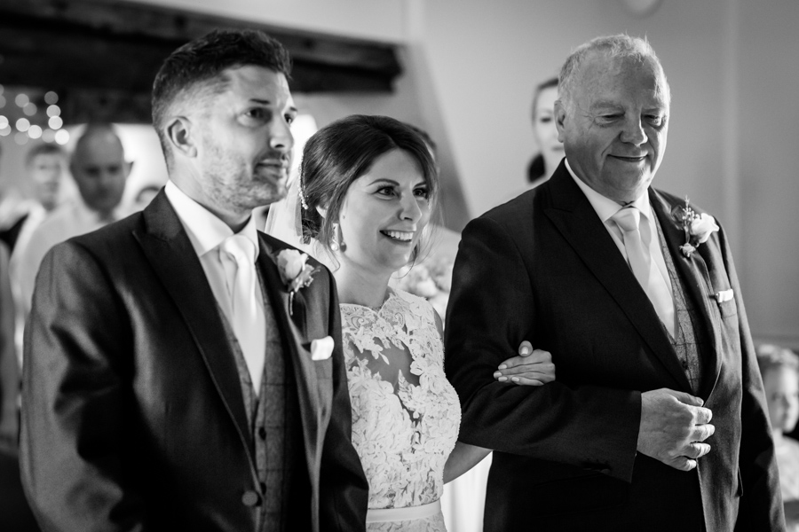 A bride smiling as she walks down the aisle accompanied by two men in suits, possibly her father and another family member or friend.