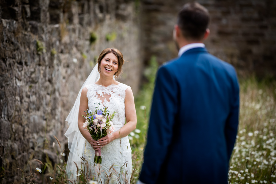 Bride smiling at groom in an outdoor setting with historic stone wall in the background.