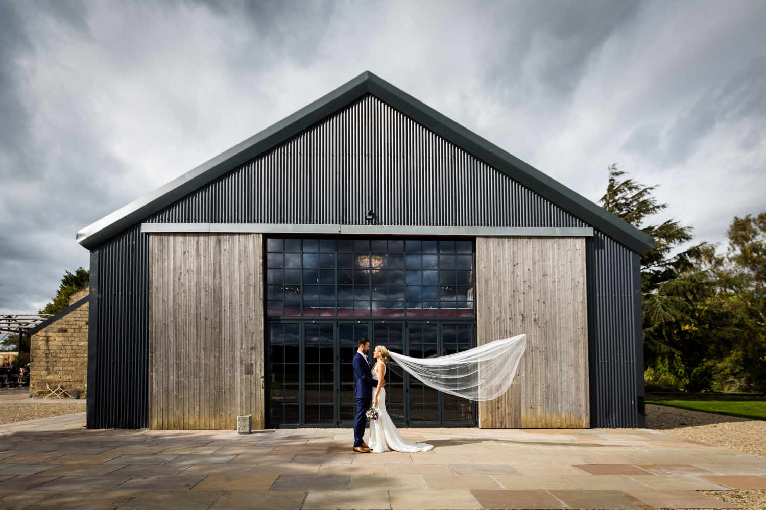 A couple in wedding attire sharing an intimate moment in front of a modern barn-style building with a dramatic cloudy sky overhead.