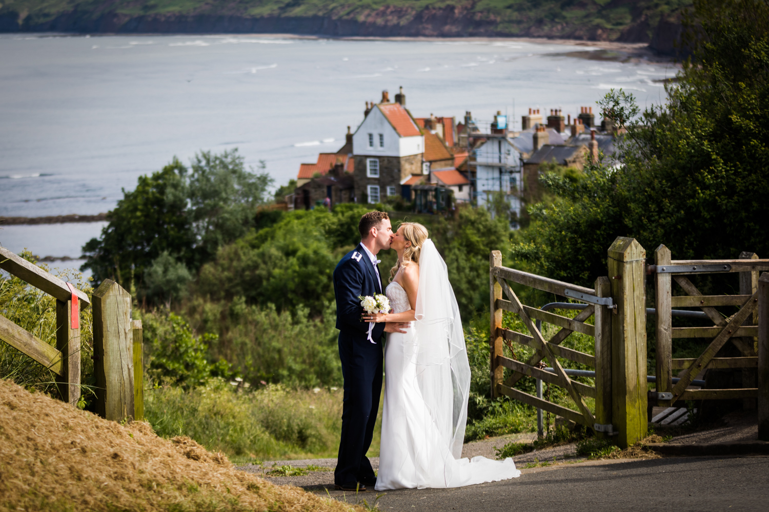 A bride and groom sharing an intimate moment on a rural path with a coastal village in the background.