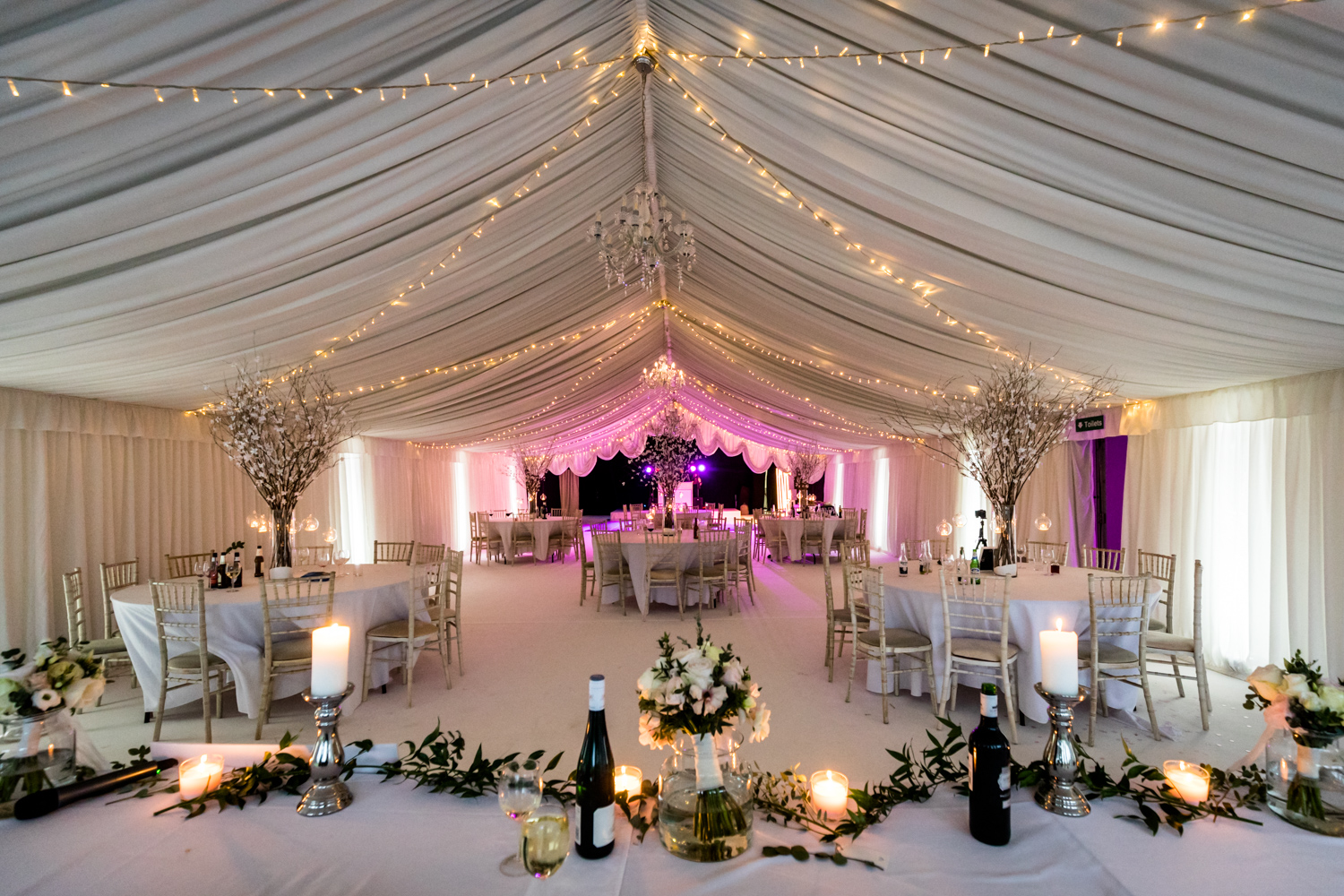 Elegant wedding reception venue decorated with drapery, string lights, floral centerpieces, and a soft pink lighting scheme.
