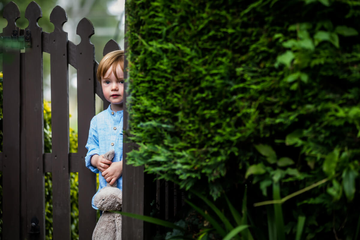 A young child clutching a stuffed toy peeks from behind a garden gate.
