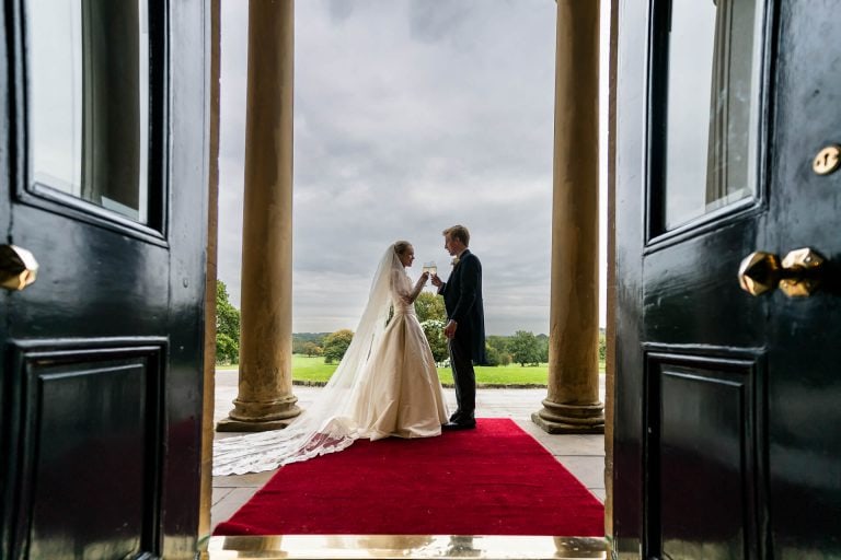 Bride and groom holding hands at the entrance of a venue, with a view of the grounds beyond.