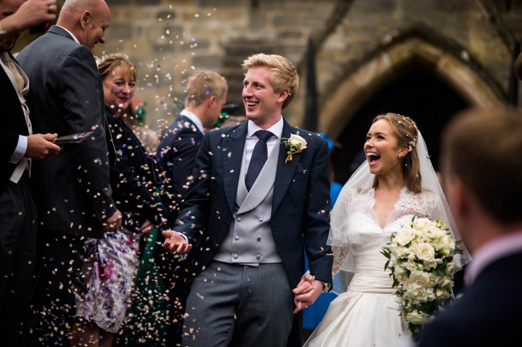 bride and groom laugh whilst guests throw confetti outside Goldsborough church