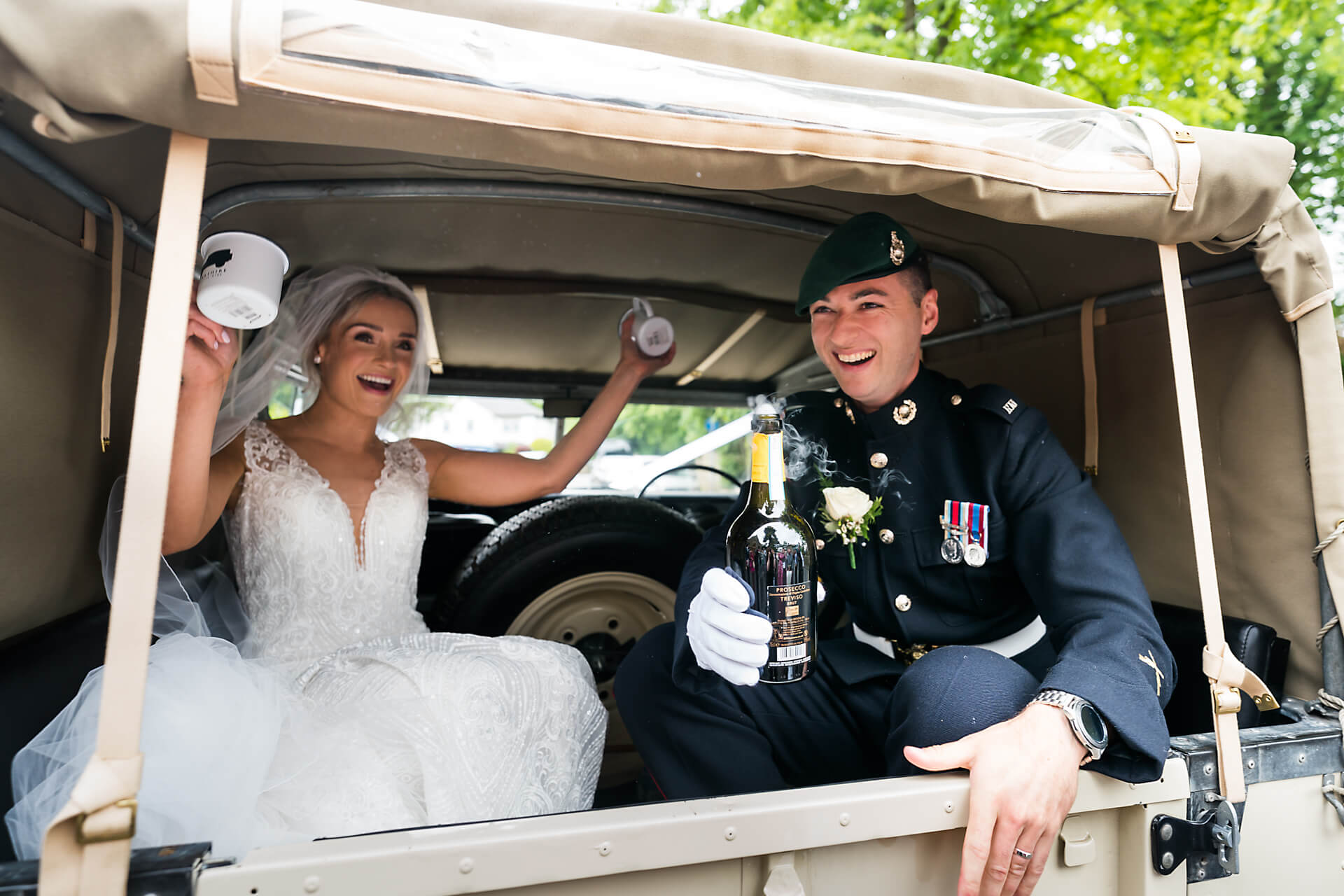 bride and groom cheer whilst holding a bottle of champagne, in the back of a landrover