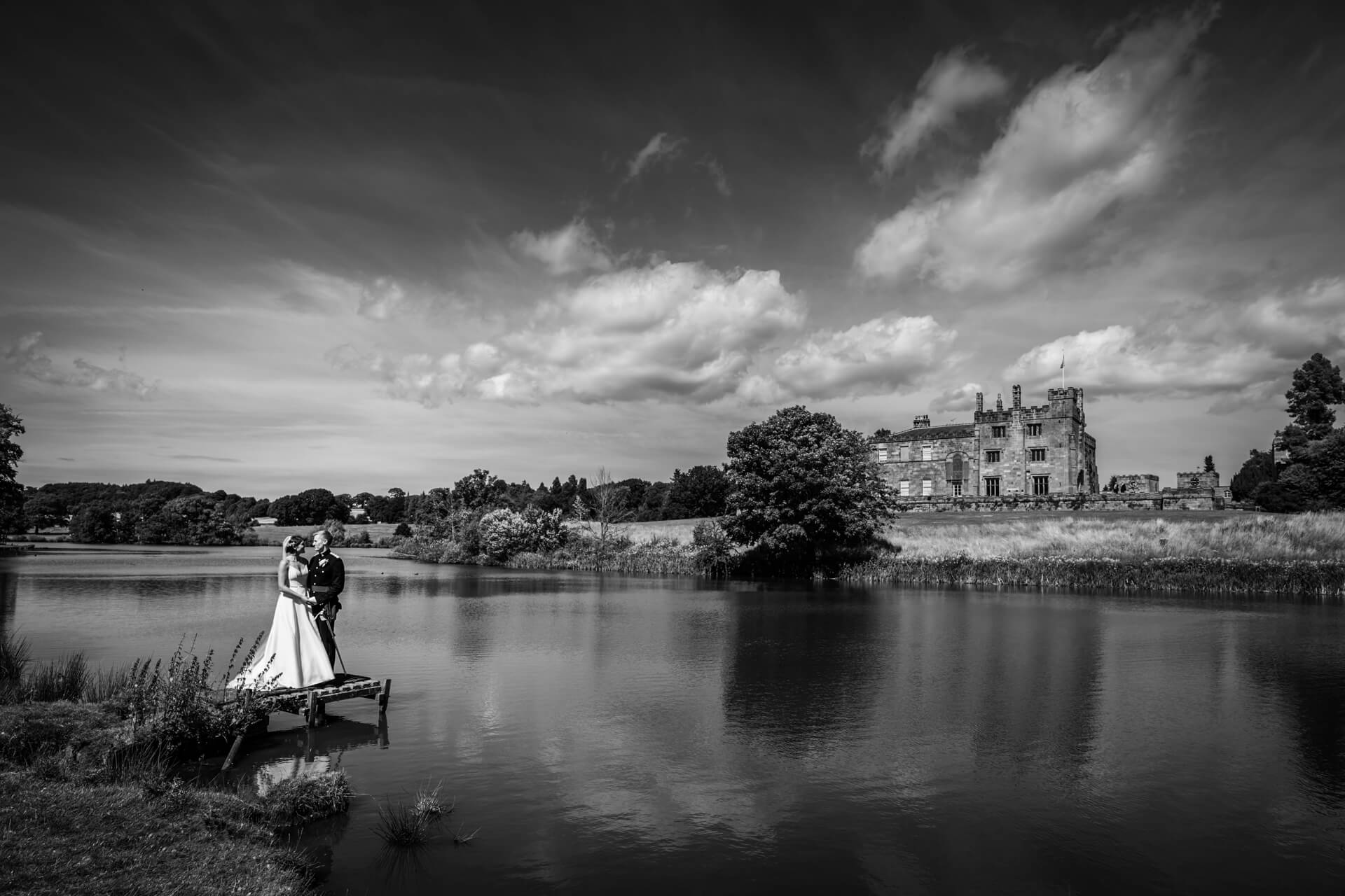bride and groom stand together next to a lake in front of Ripley Castle