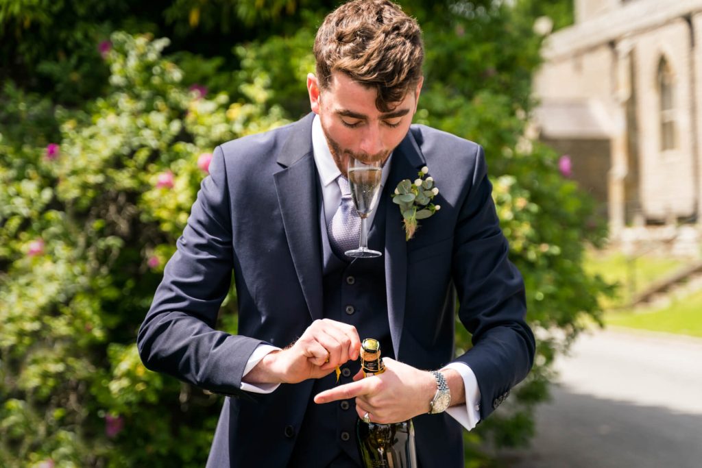 best man trying to open a champagne bottle whilst holding a glass in his mouth