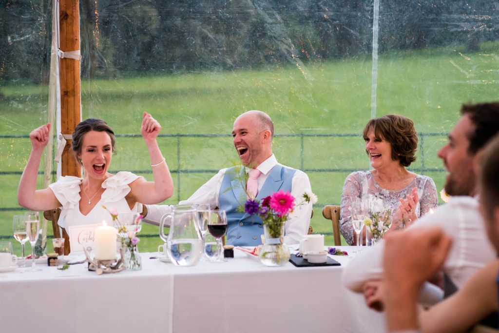 top table laughing whilst bride celebrated with her arms in the air