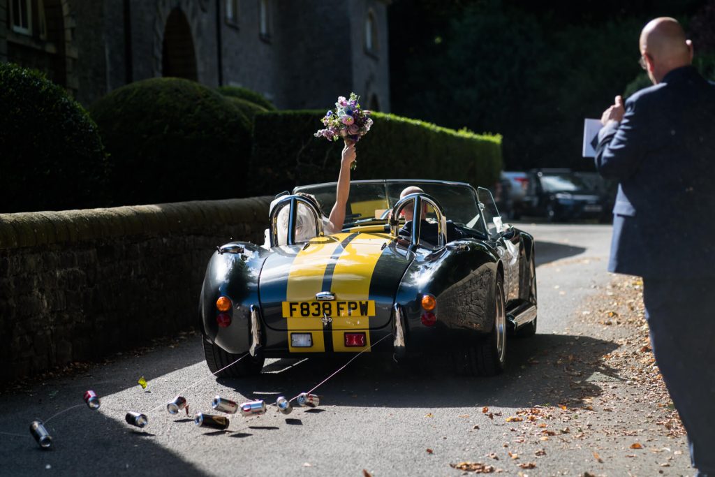 bride waves her flowers in the air as they drive through the Broughton Hall Estate