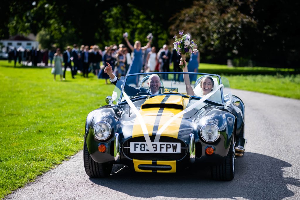 wedding couple cheer from their car as they drive through the Broughton Hall Estate