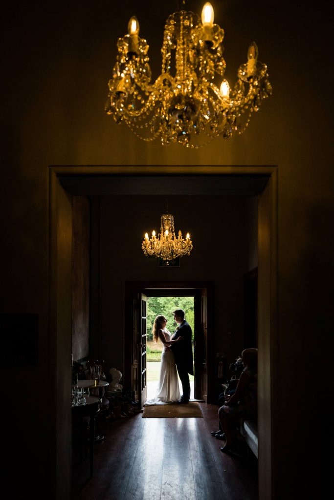 bride and groom hugging in a doorway under some chandeliers at Thicket Priory