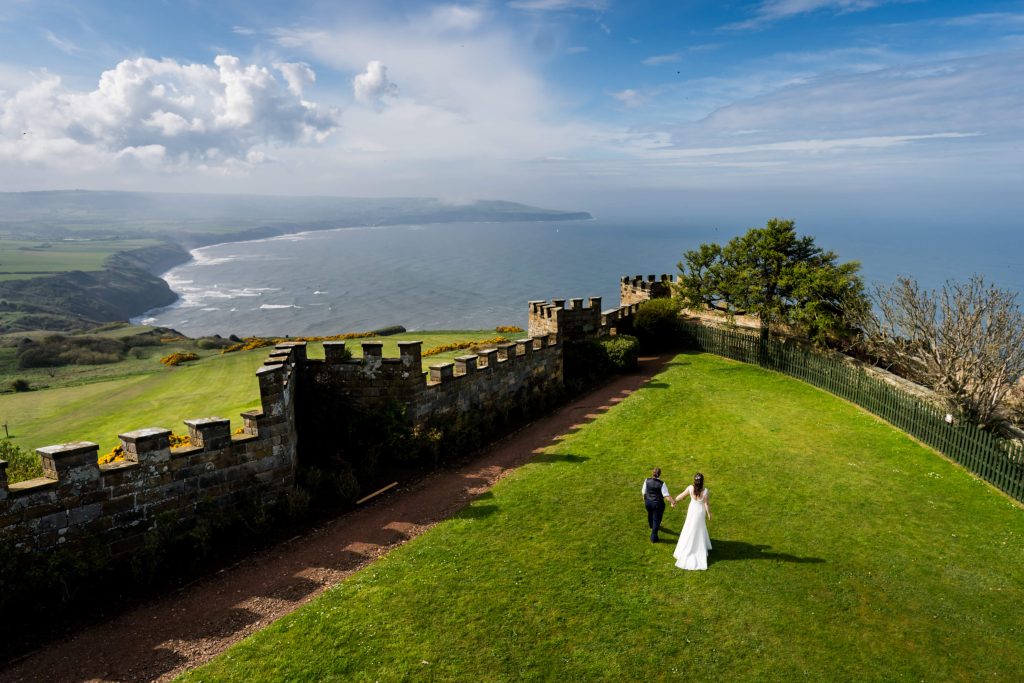 Wedding couple by coastal Raven Hall castle ruins in sunlight.