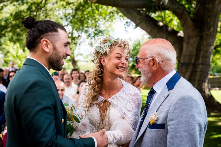 Bride, groom, and the bride's father smiling at outdoor wedding.