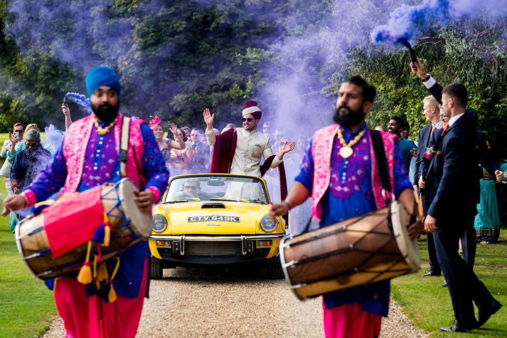 Festive Indian wedding procession with traditional drummers and classic car.