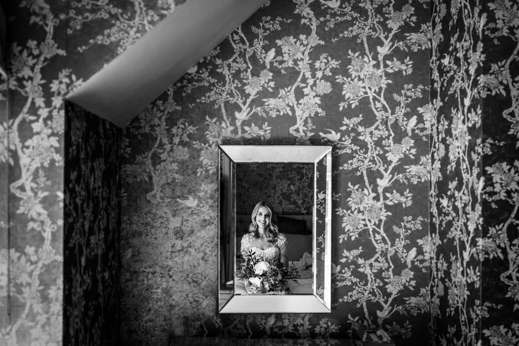 Smiling bride in framed mirror, floral wallpaper, black and white.