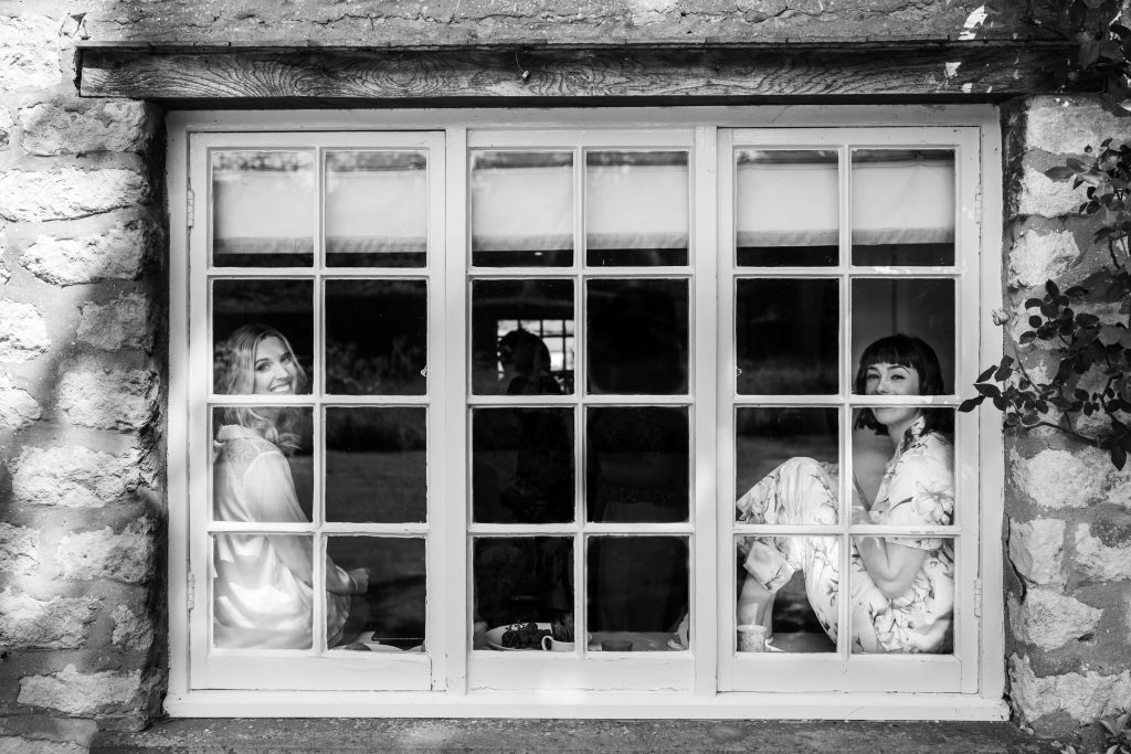 Bride and bridesmaid smiling behind windowpane in black and white.