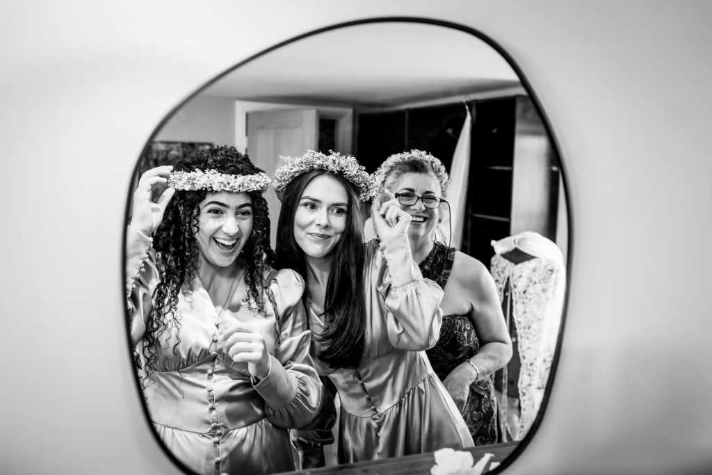 Three women smiling, reflected in mirror, wearing floral crowns.