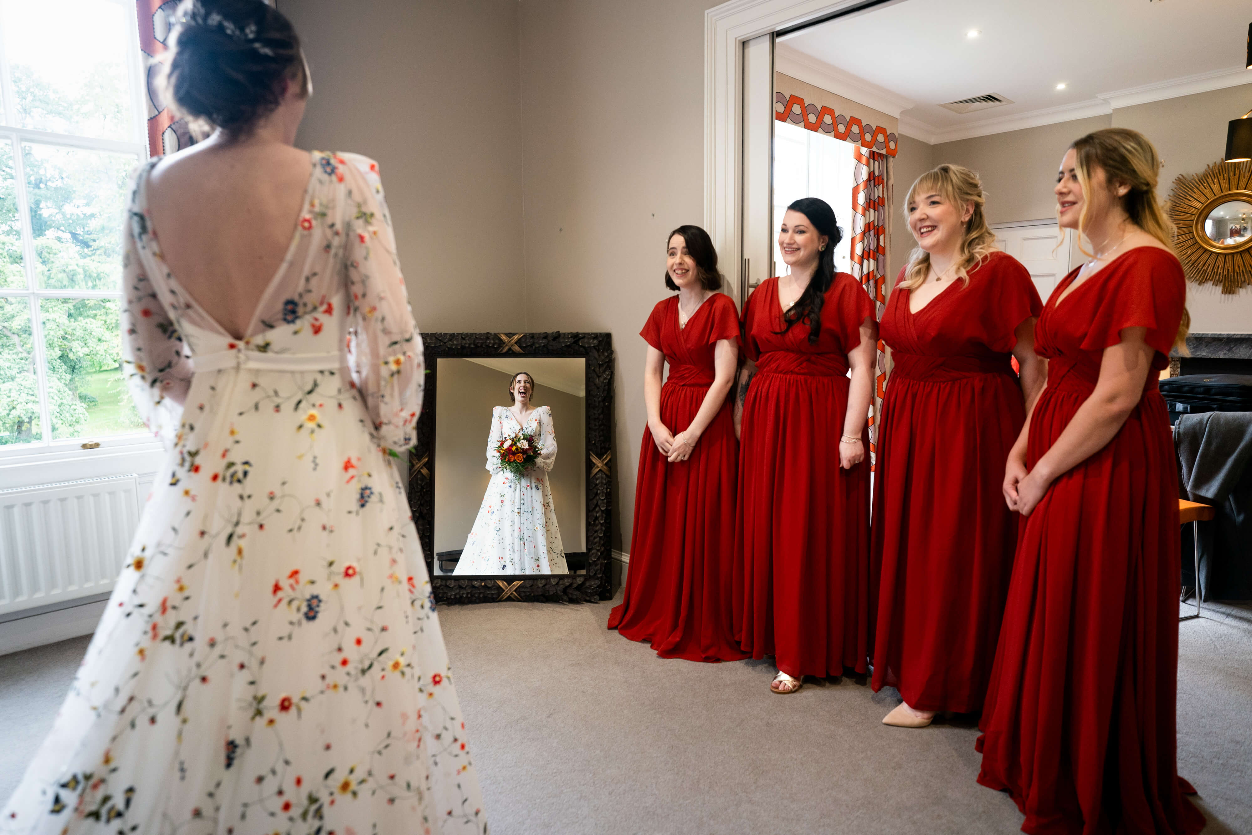 Bride reflection in mirror with smiling bridesmaids in red dresses.