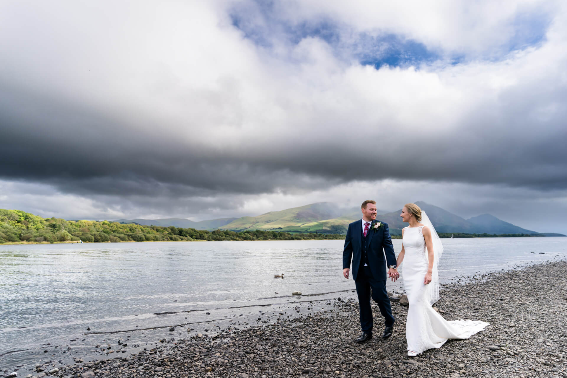 A bride and groom walking along a pebble beach with a scenic view of mountains and a cloudy sky.