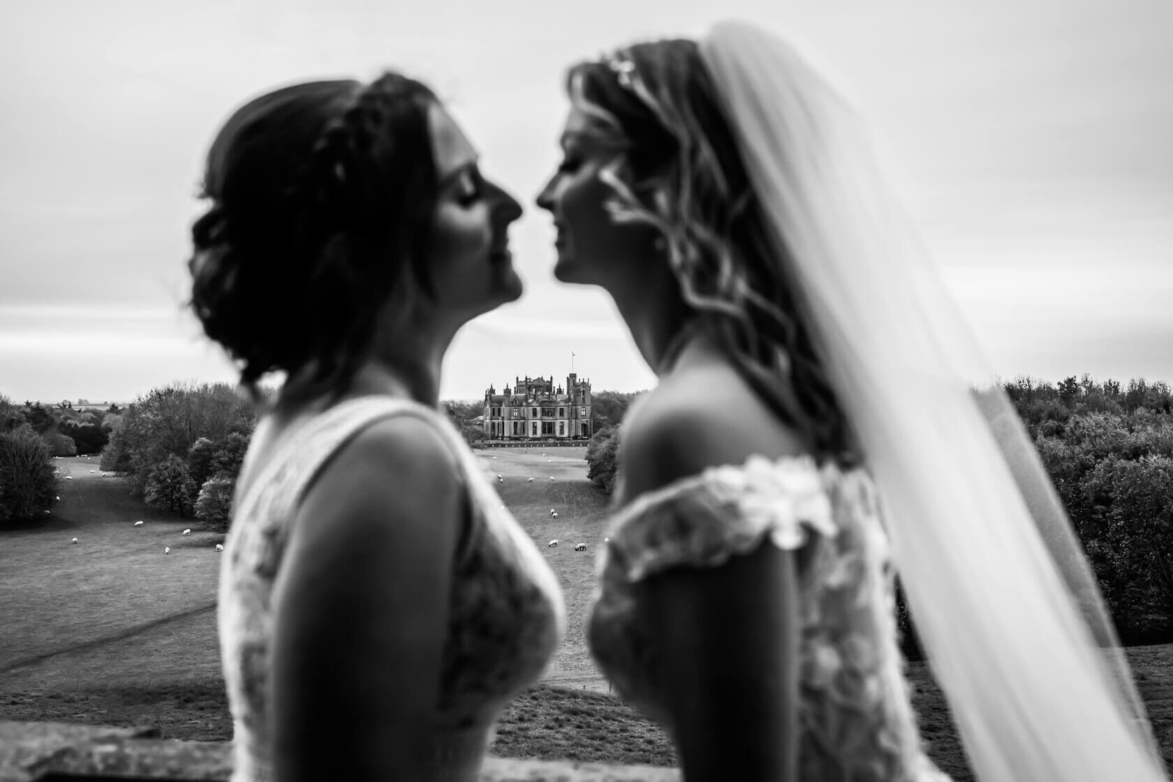 Two brides face each other in black and white with Allerton Castle in the distance