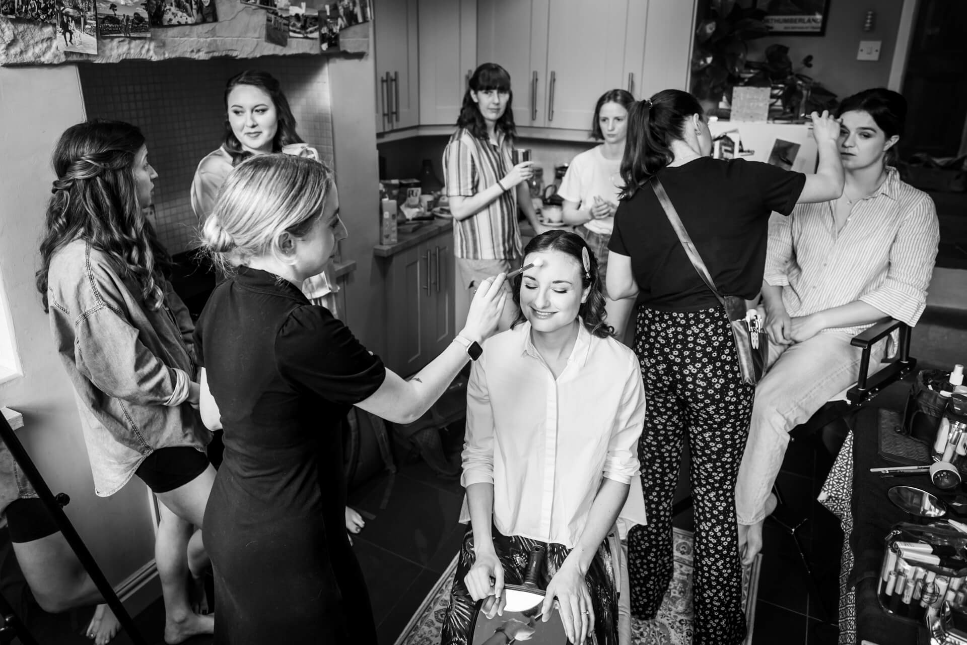Bride getting makeup done, friends around in kitchen.