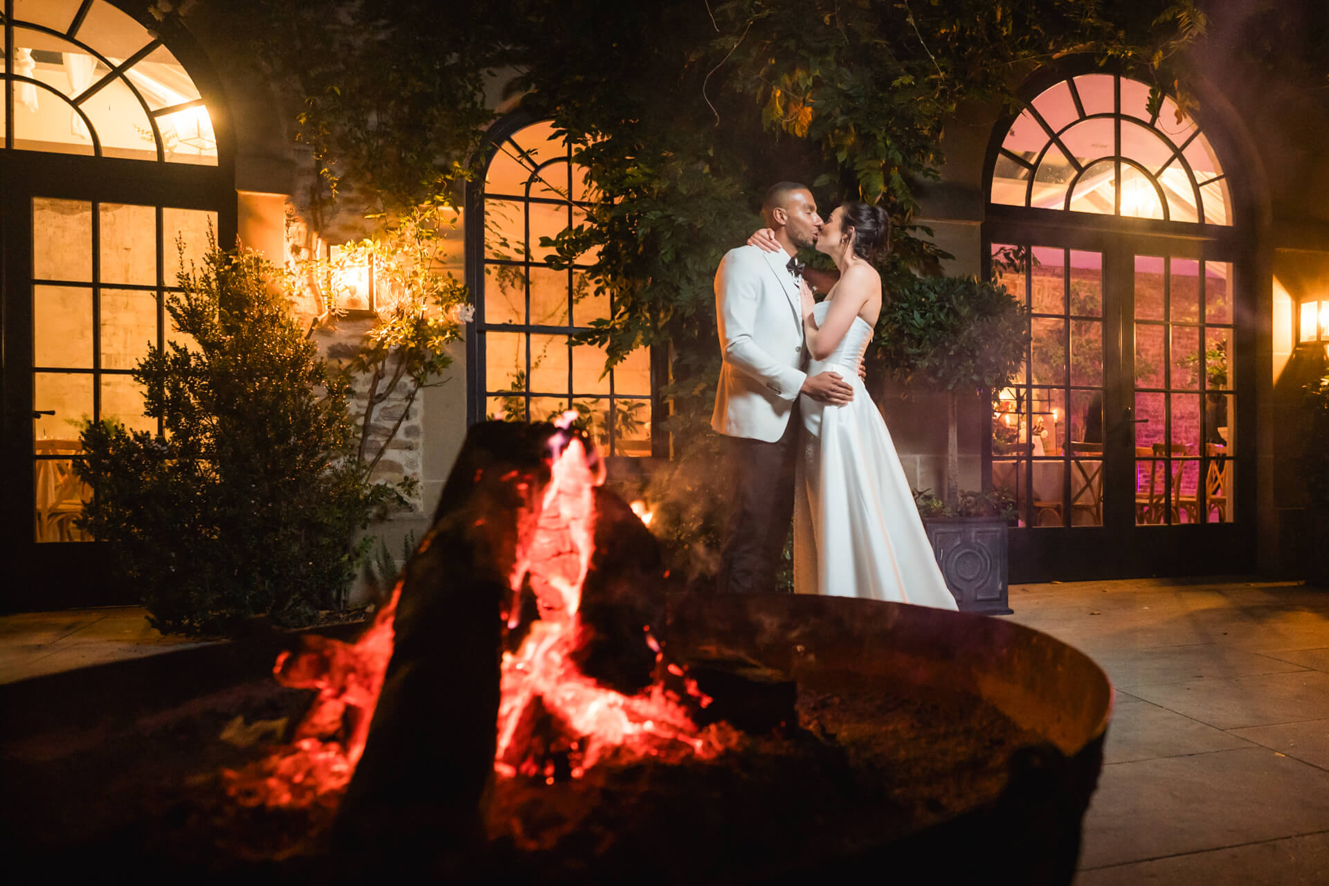 Couple kissing by fire outside elegant venue.