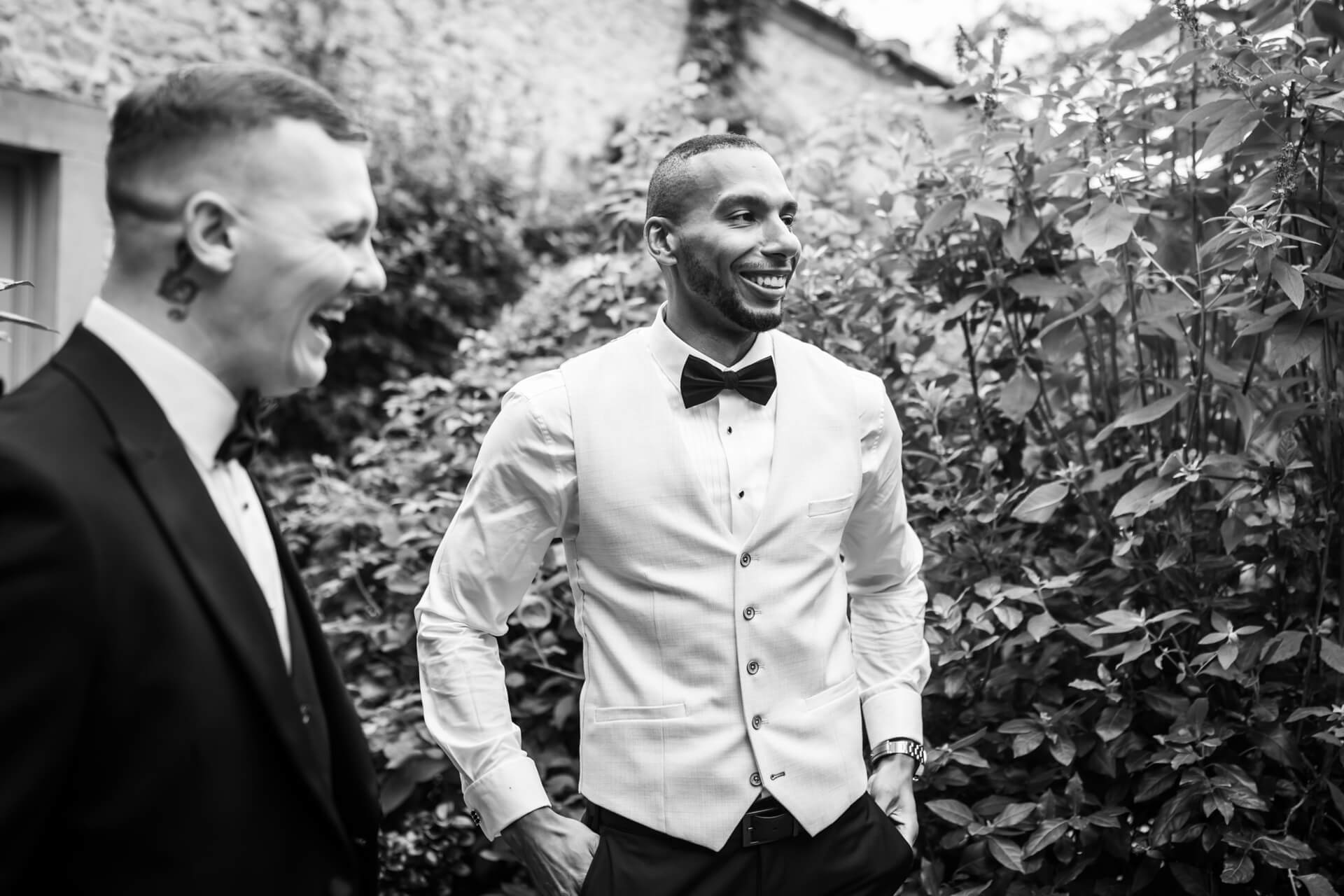 Two groomsmen laughing in garden, wearing suits.