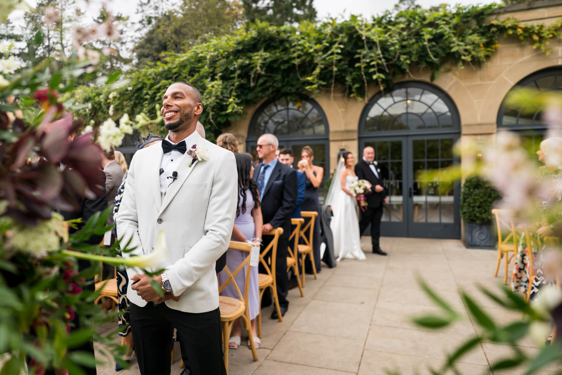 Groom smiles at outdoor wedding ceremony, guests watching.