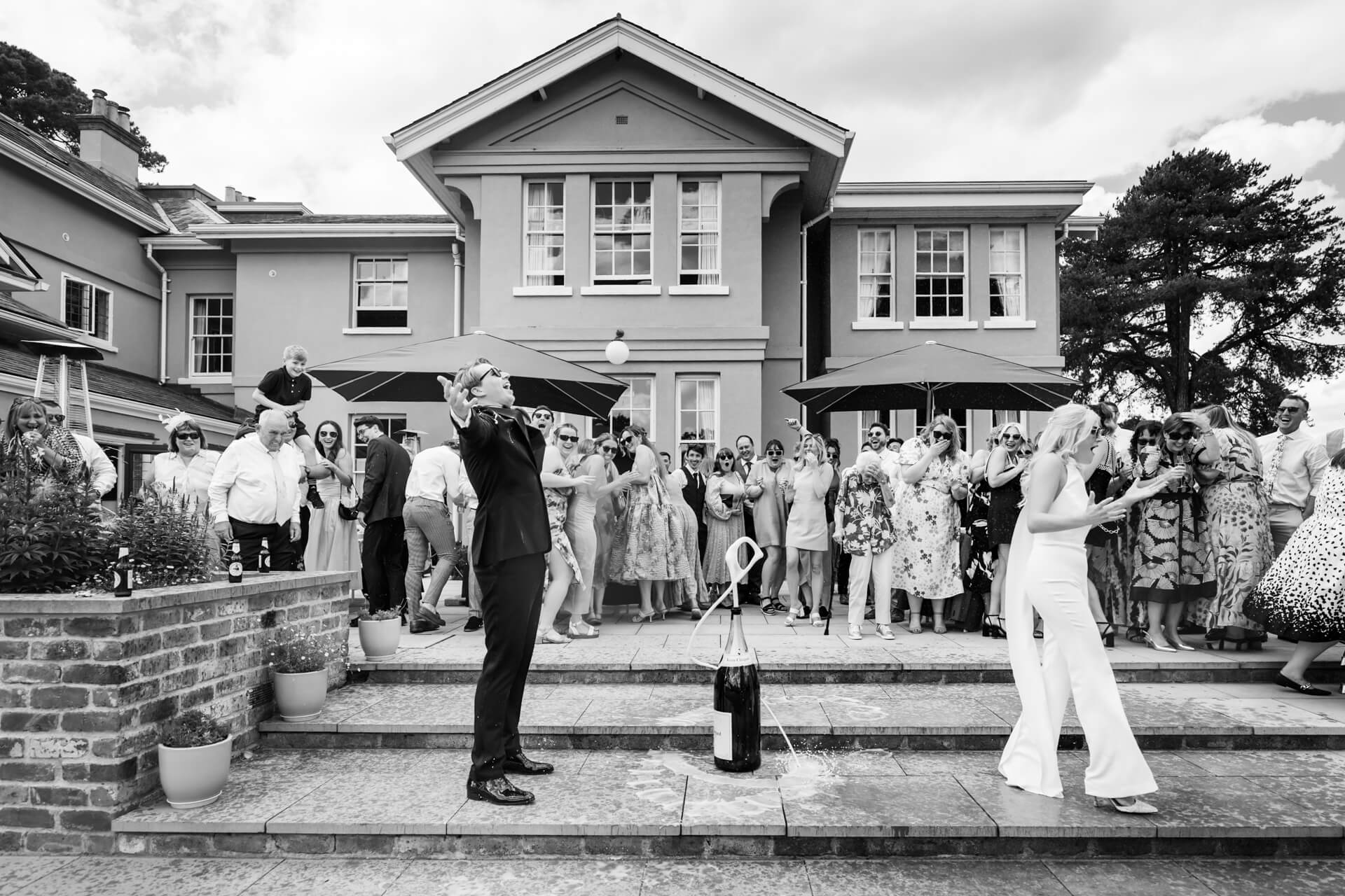 The wedding guests standing on a patio, reacting to a large champagne bottle spraying foam in front of a large house.