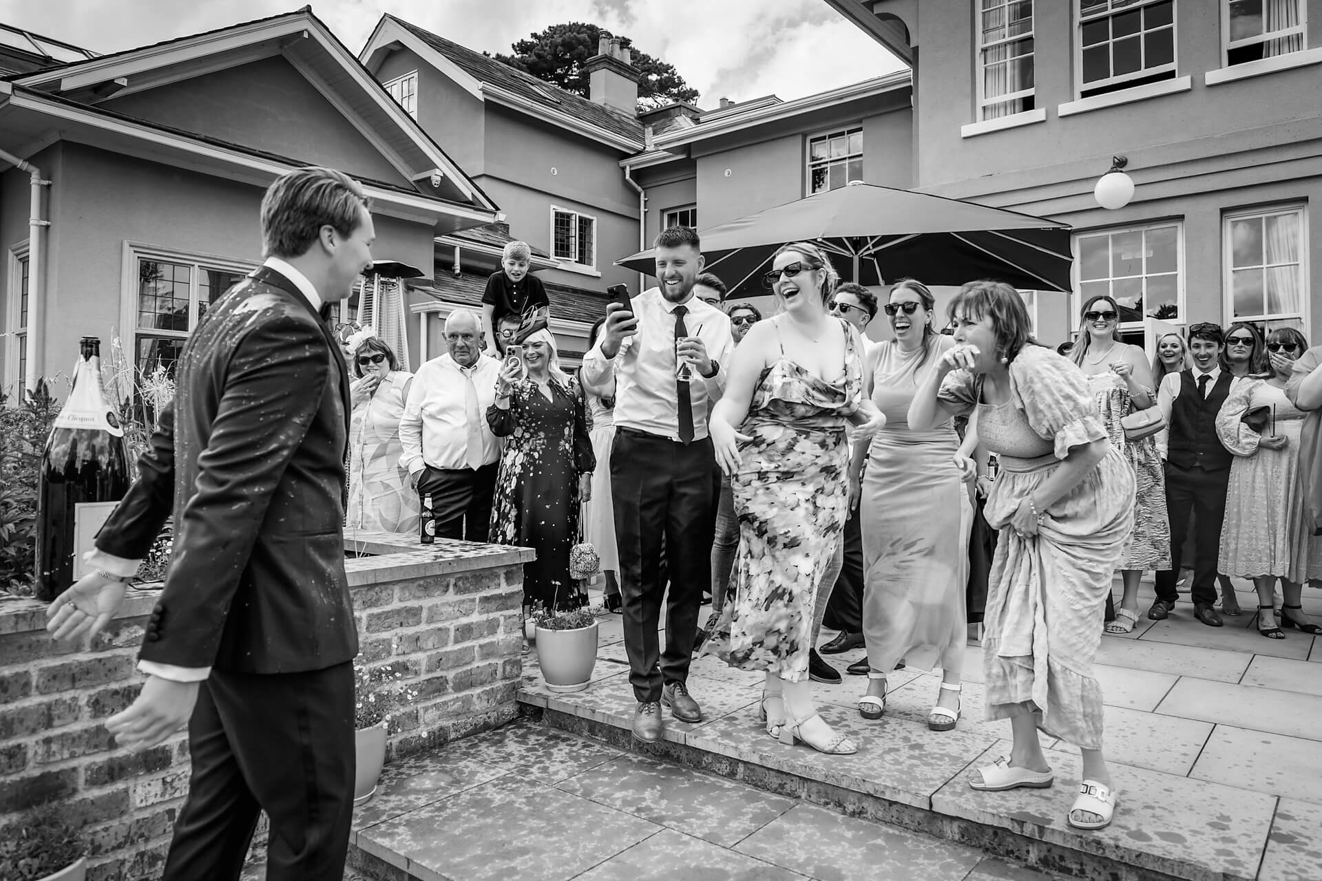 A group of guests outdoors at a wedding react with surprise and laughter as the groom is soaked in champagne