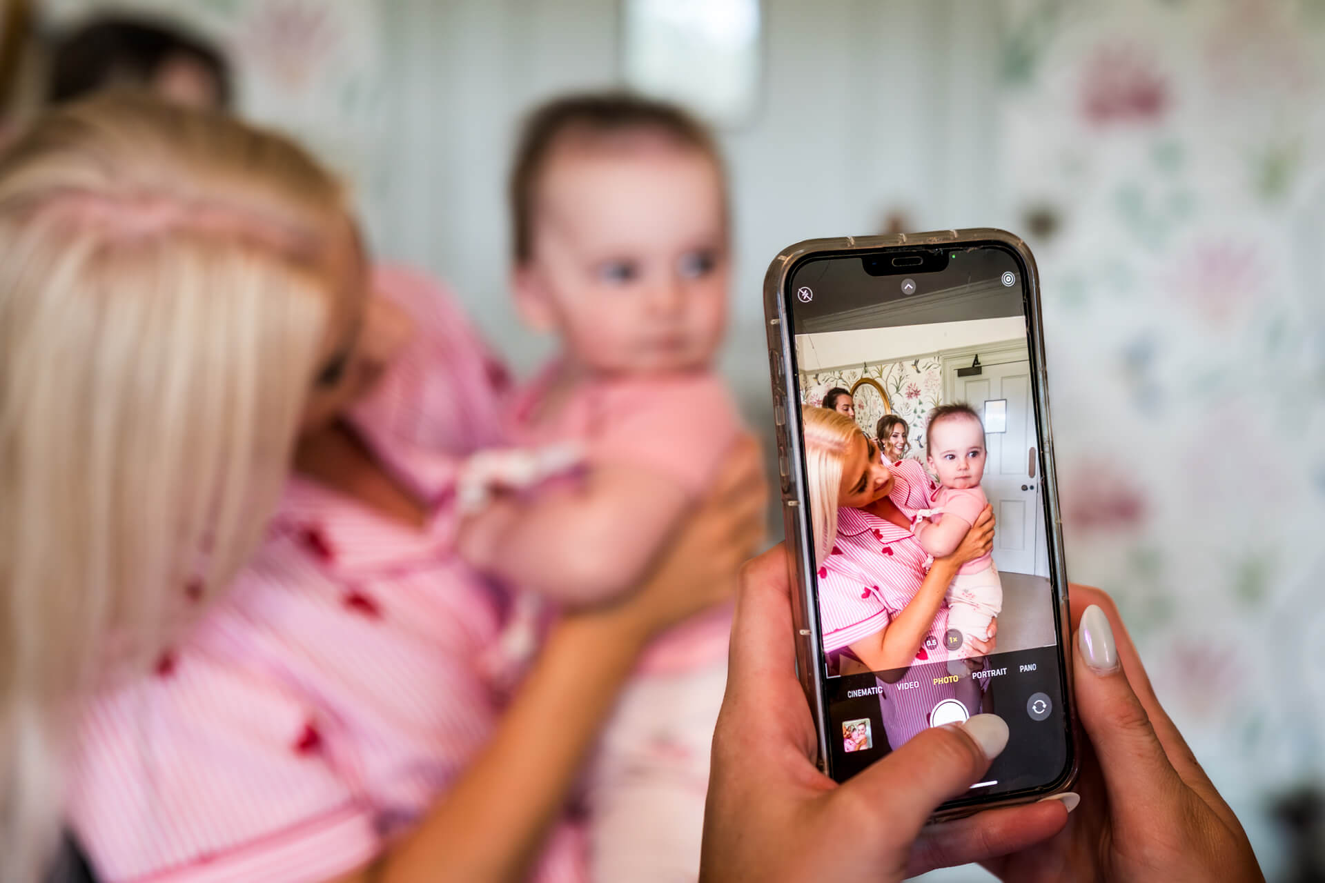 A person holds a mobile, taking a photo of an adult holding a baby. The mobile screen displays the captured image, and both wear pink clothes. Background is slightly blurred.