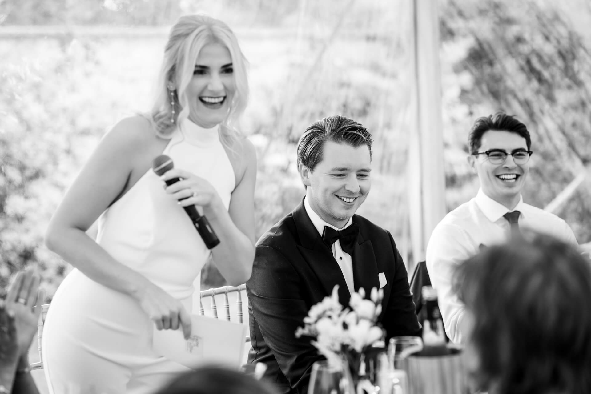 A bride speaks into a microphone beside two men in suits who are seated and smiling at a formal event.