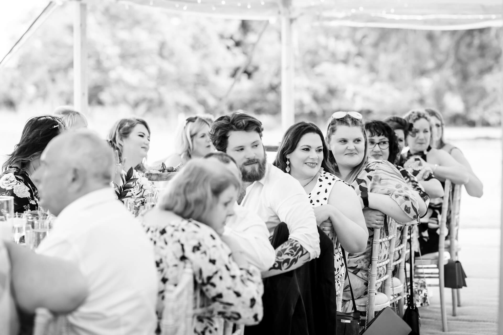 A group of people seated at tables outdoors, turning to look in the same direction, during a formal event.