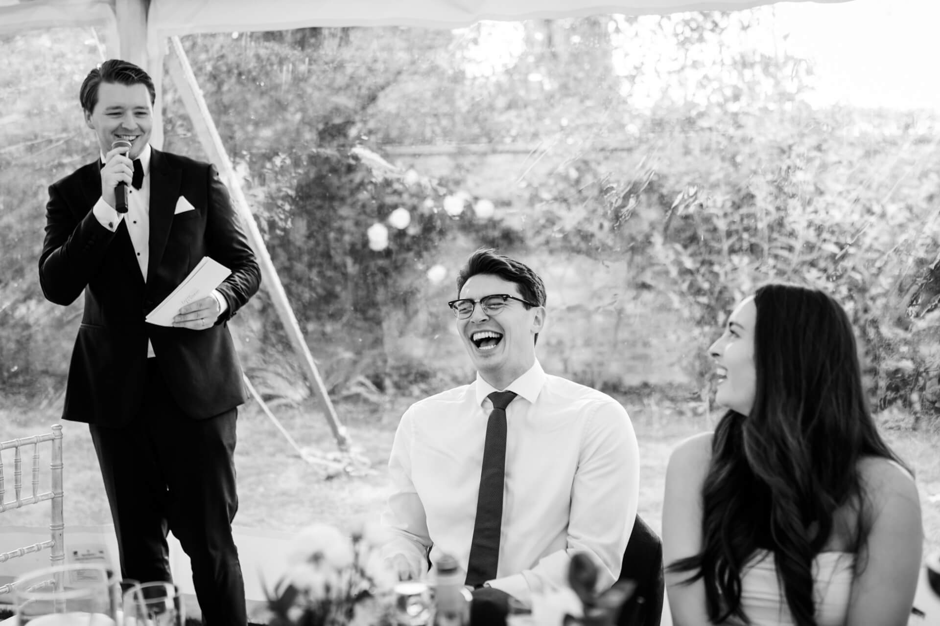 A groom in a dinner jacket speaks into a microphone while a seated man and woman laugh, outdoors at what appears to be a formal event.