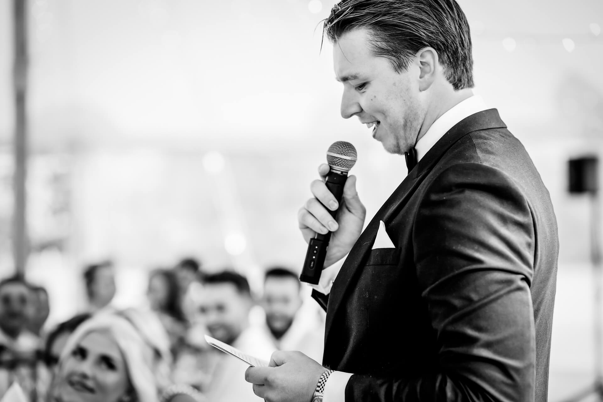 a groom holding a microphone and reading from a card, speaking to an audience at an event. The image is in black and white.