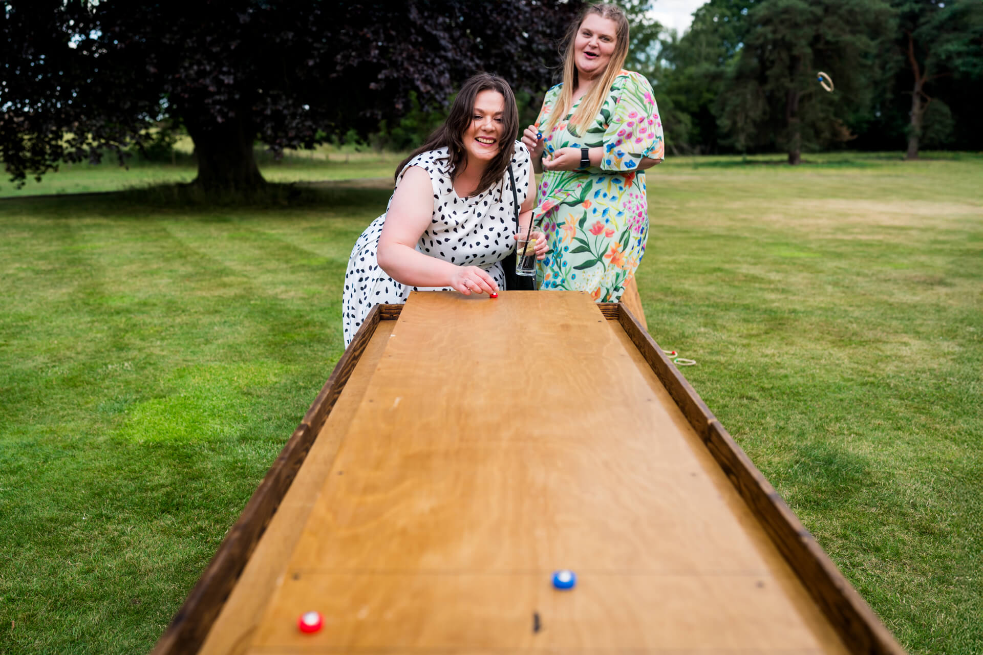 Two women, one in a white polka dot dress and one in a floral dress, play a wooden tabletop game outdoors on a grassy lawn.