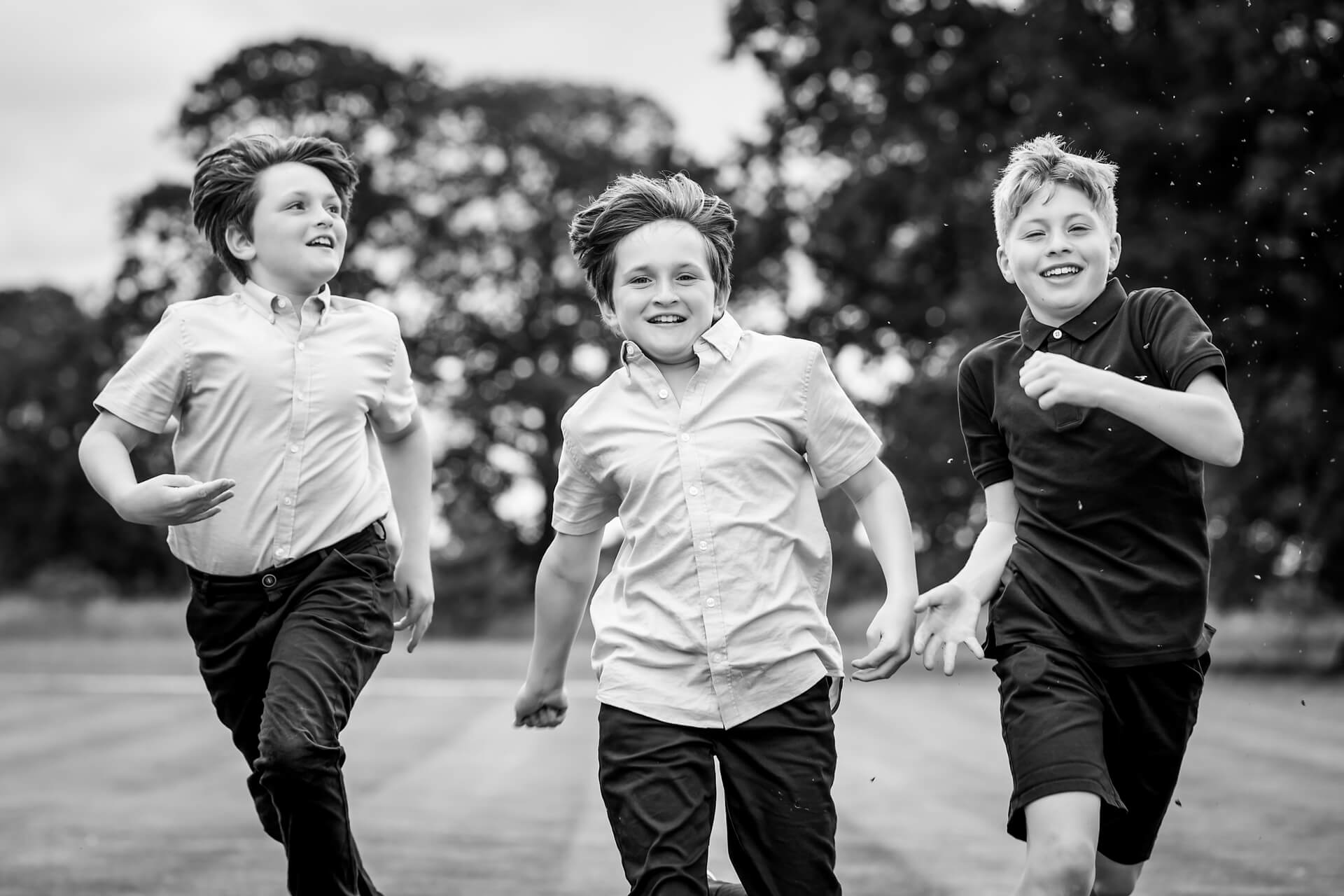Three boys run towards the camera outdoors on grass, all smiling and wearing casual shirts. Trees are visible in the blurred background. The image is in black and white.