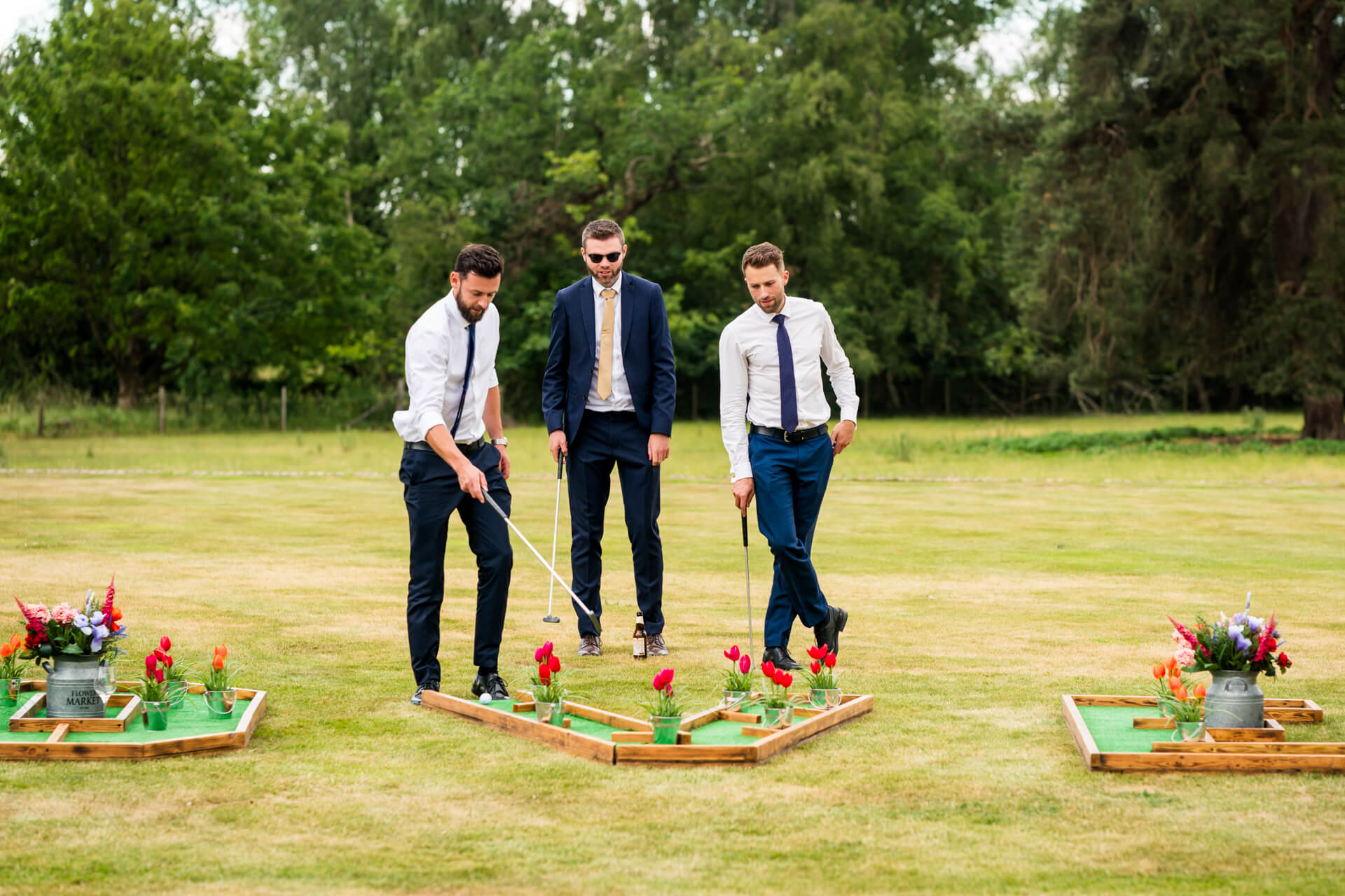 Three men in formal attire play crazy golf outdoors on a grassy lawn, with flower decorations around the crazy golf holes.