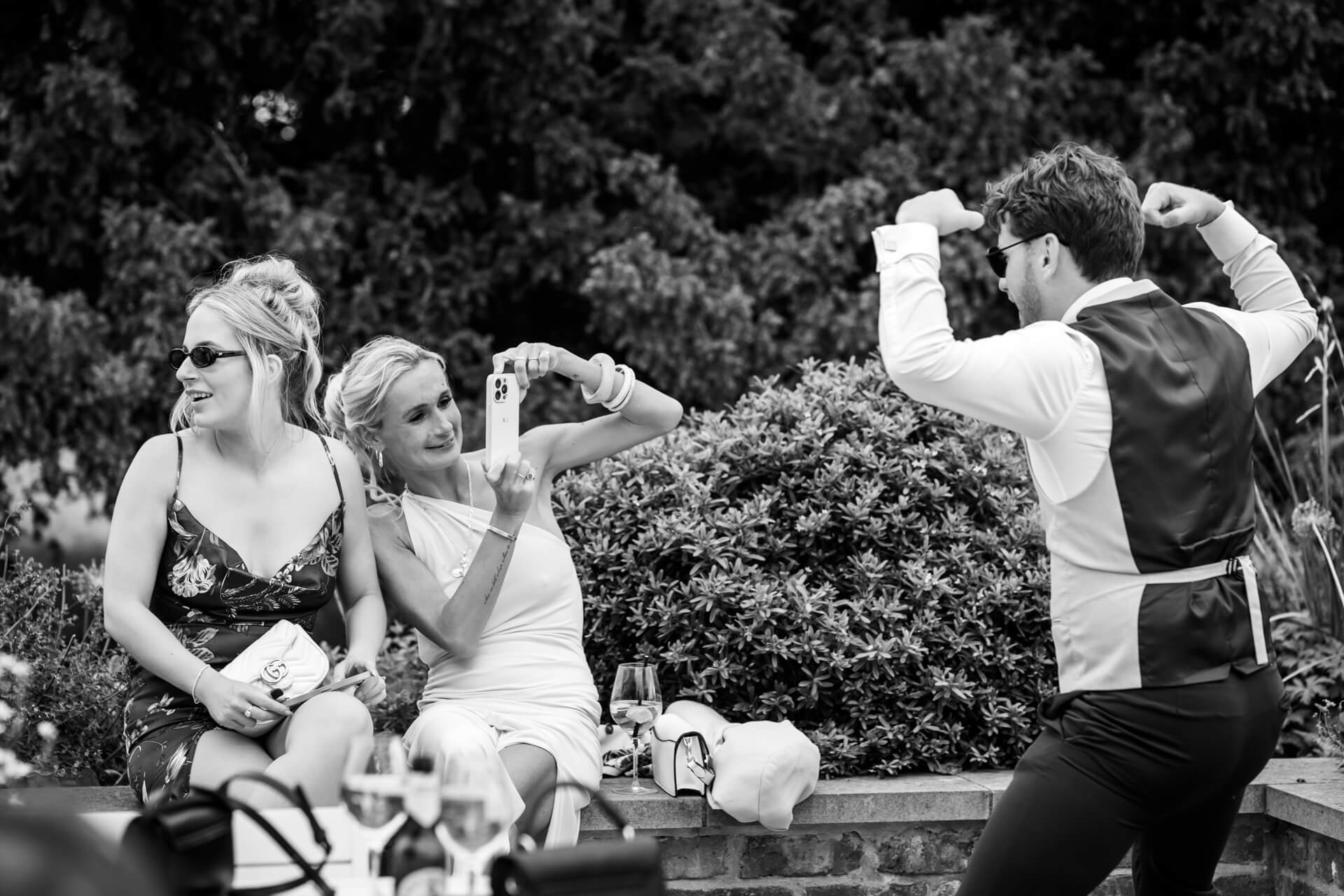 Two women sit on a low wall, one taking a photo with her mobile, while a man in a waistcoat flexes his arms and poses in front of them. The setting appears to be outdoors.