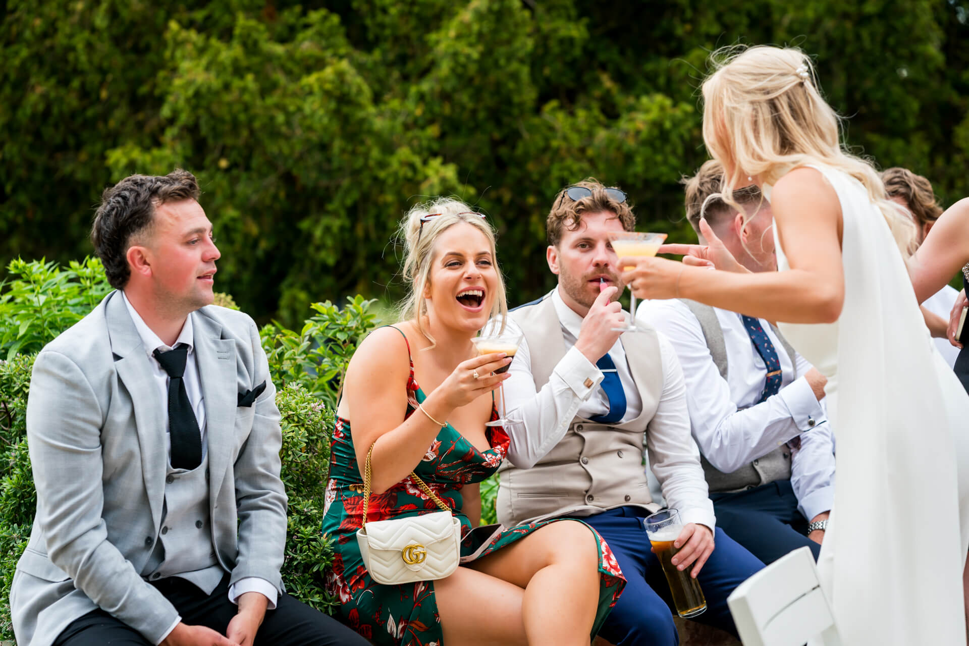 A group of people dressed in formal attire sit and stand outdoors, holding drinks and talking, with greenery in the background.