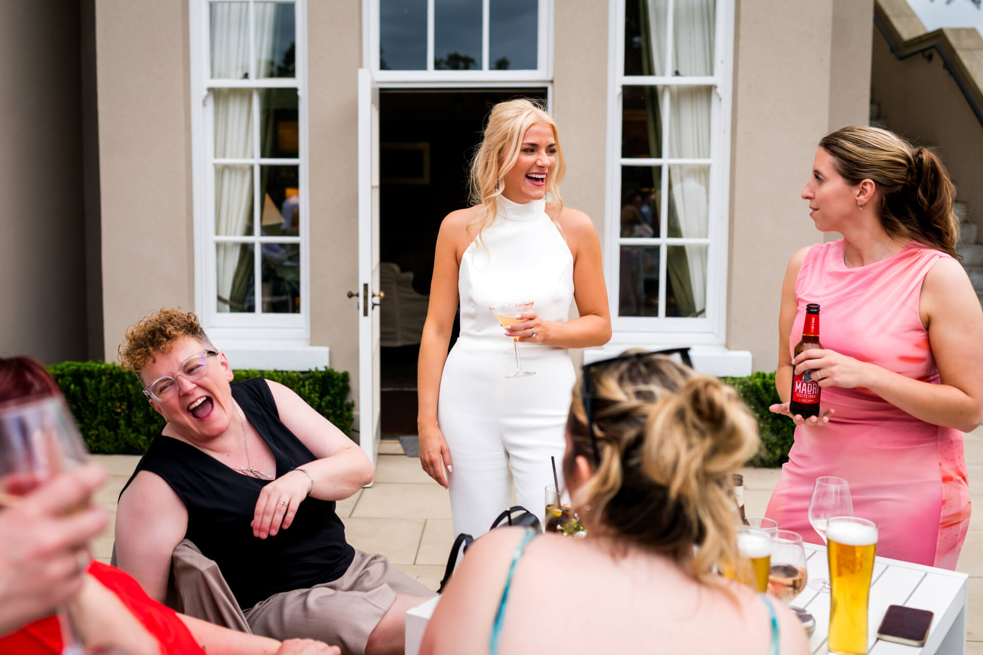 A bride stands and smiles with a drink in hand, surrounded by others sitting and laughing outside a building.