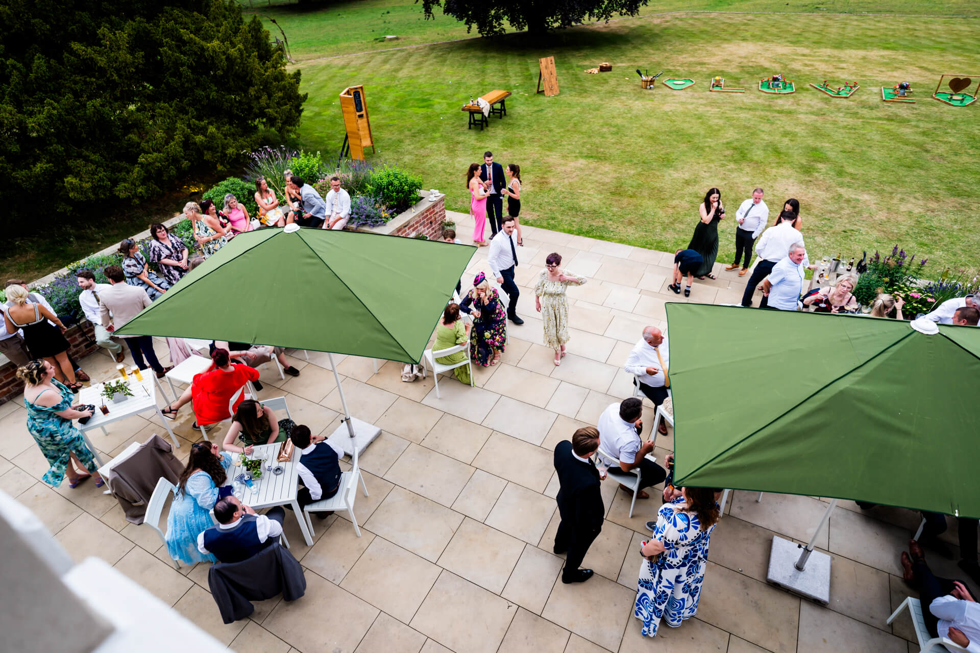 An outdoor gathering with people socialising under large green umbrellas on a patio, with lawn games set up on the grass in the background.