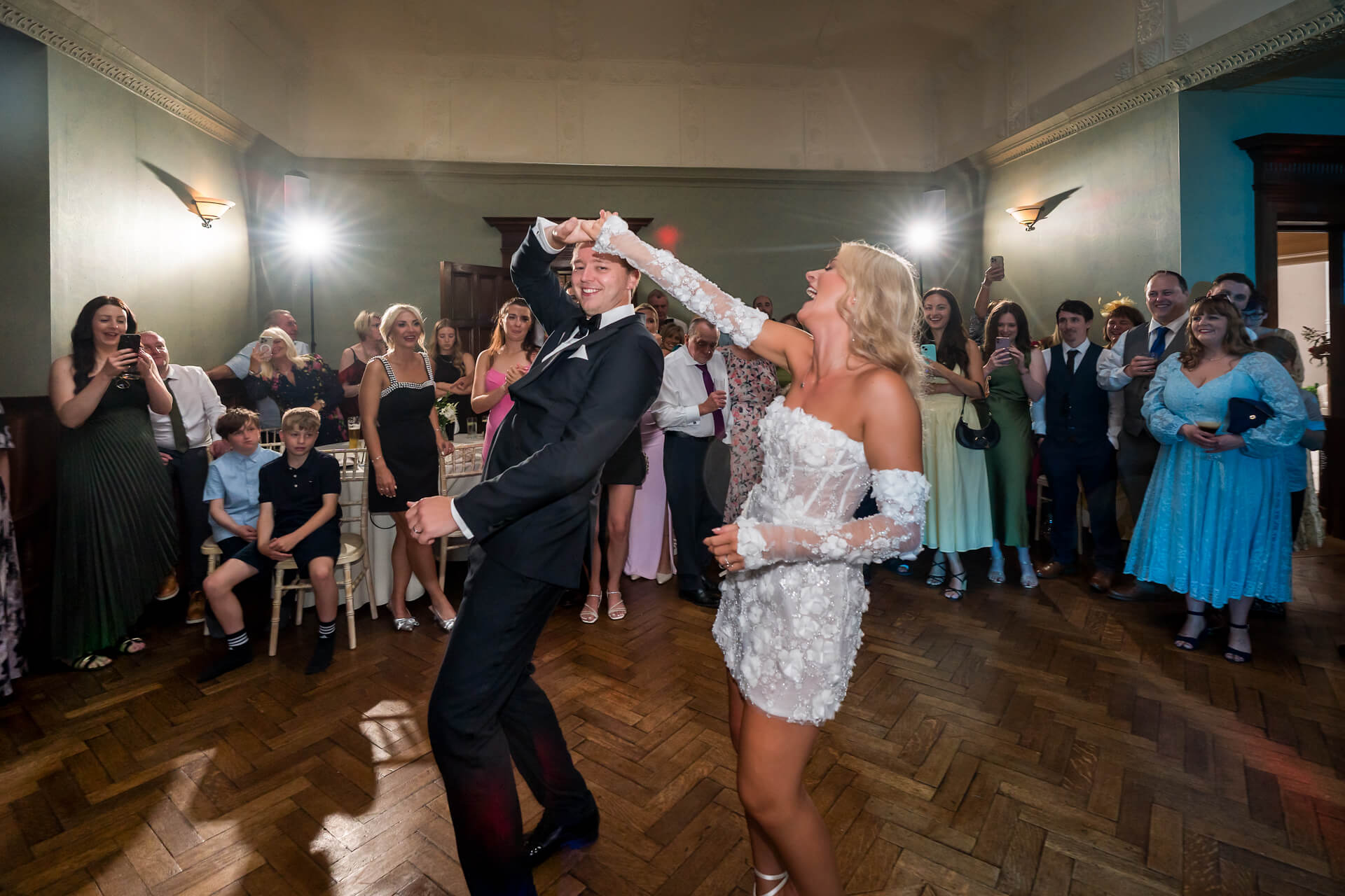 A bride and groom dance together in the centre of a room while guests watch and smile, some taking photos; the bride wears a short white dress, and the groom wears a black suit.