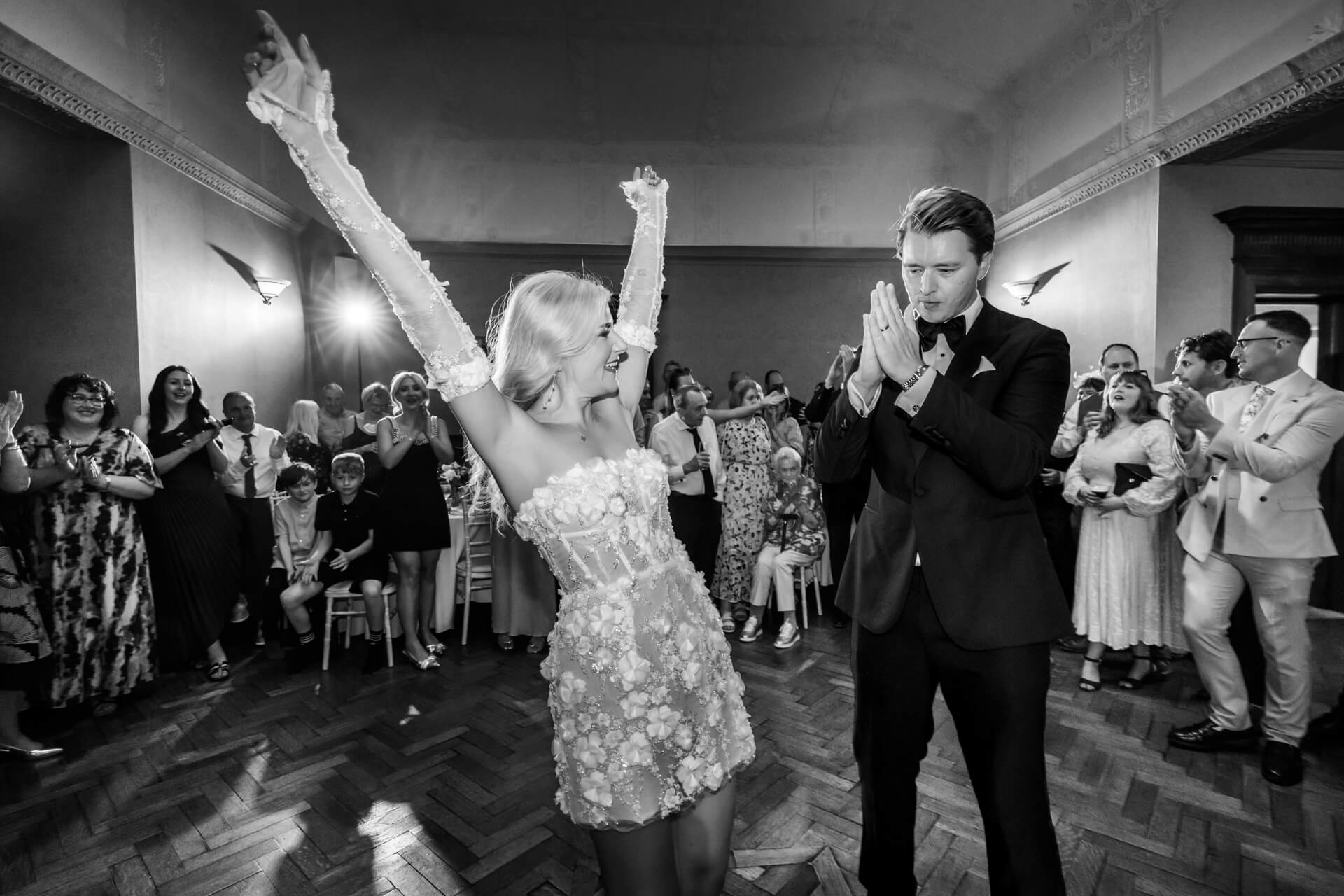 A bride with arms raised in celebration faces a groom who clasps his hands, surrounded by applauding wedding guests in a decorated hall.