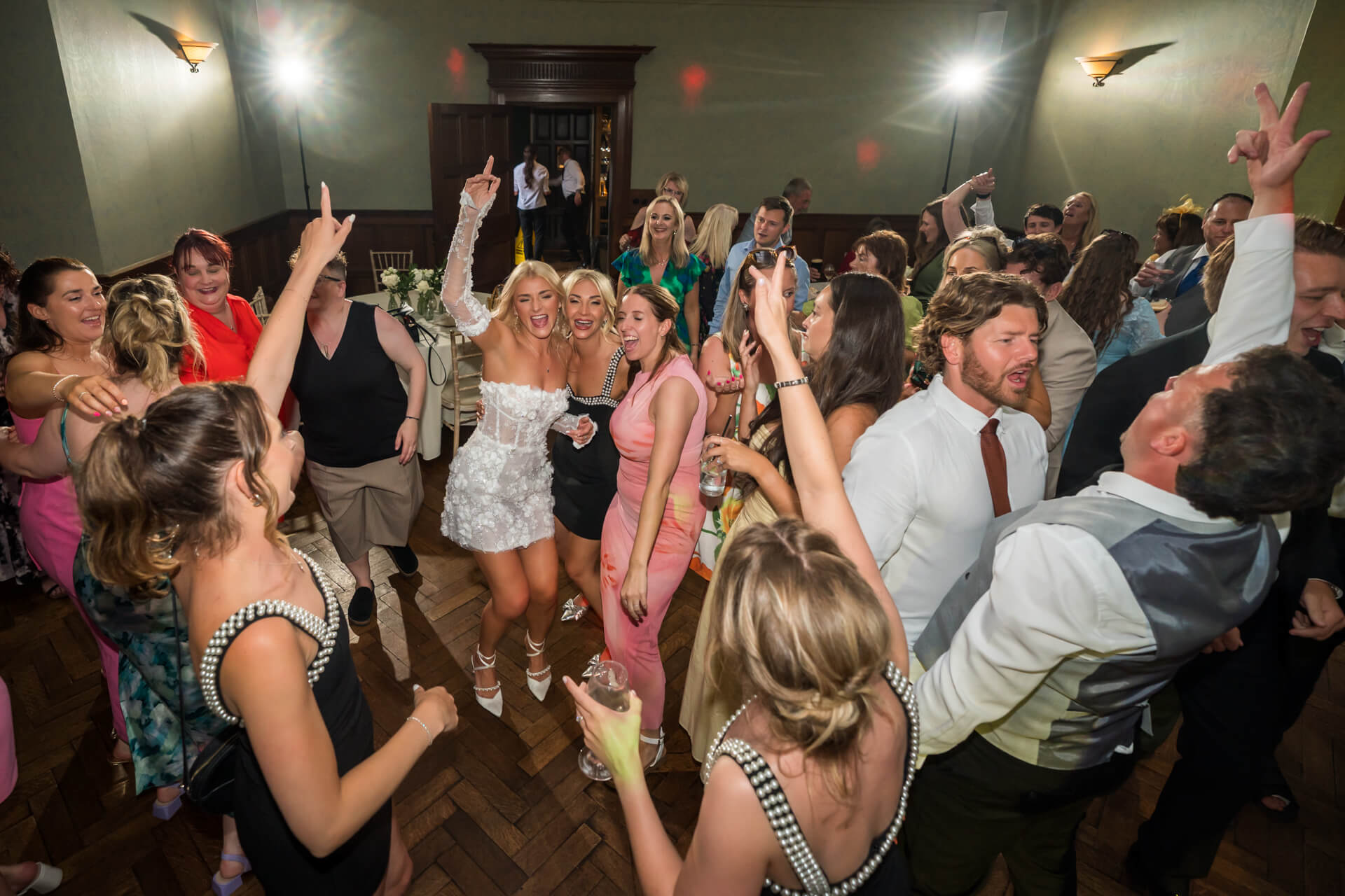 A group of people dance and celebrate indoors at a party or wedding, with guests smiling and raising their arms in excitement.