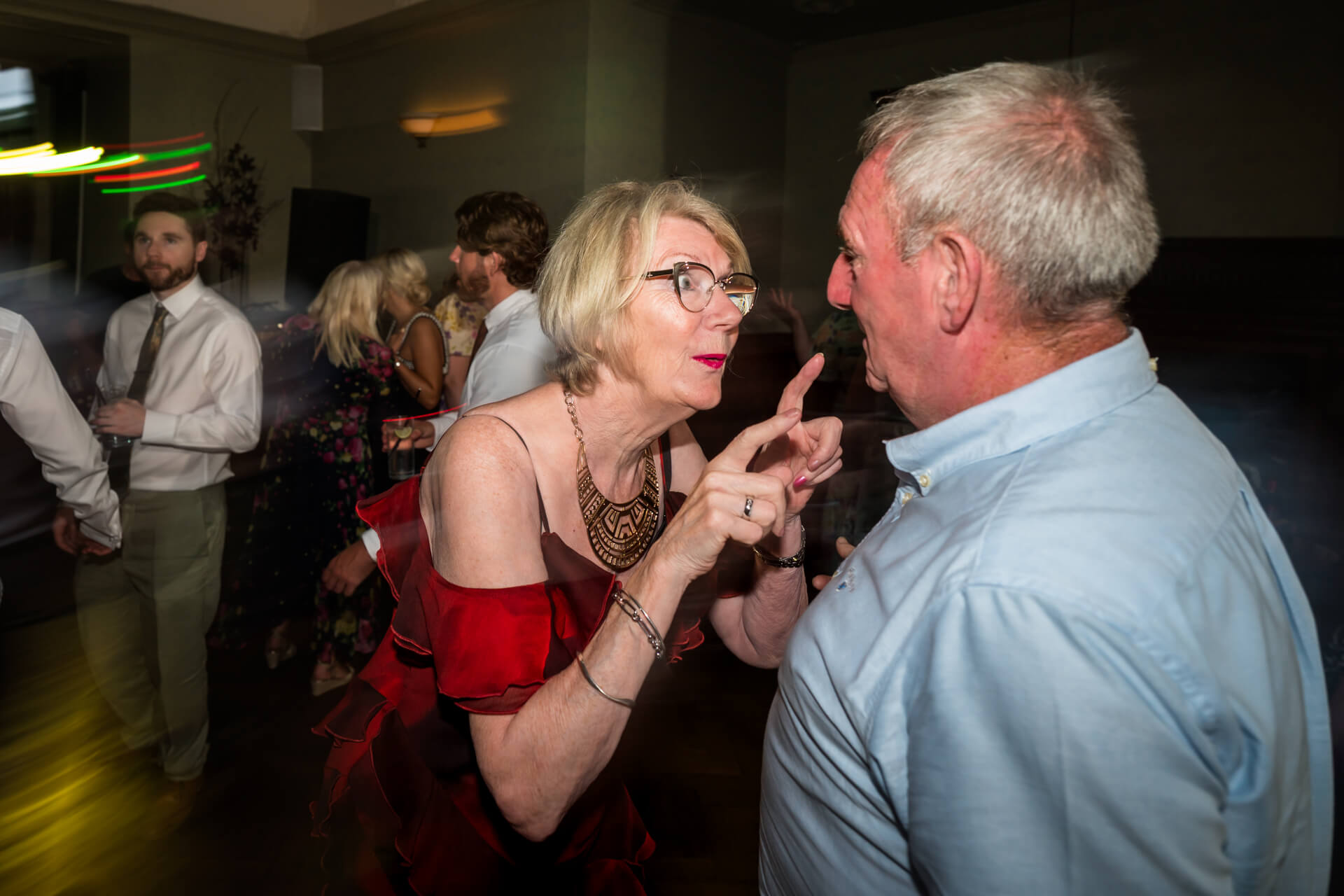 An older woman in a red dress gestures and speaks to an older man in a light blue shirt at a party, with other people dancing in the background.