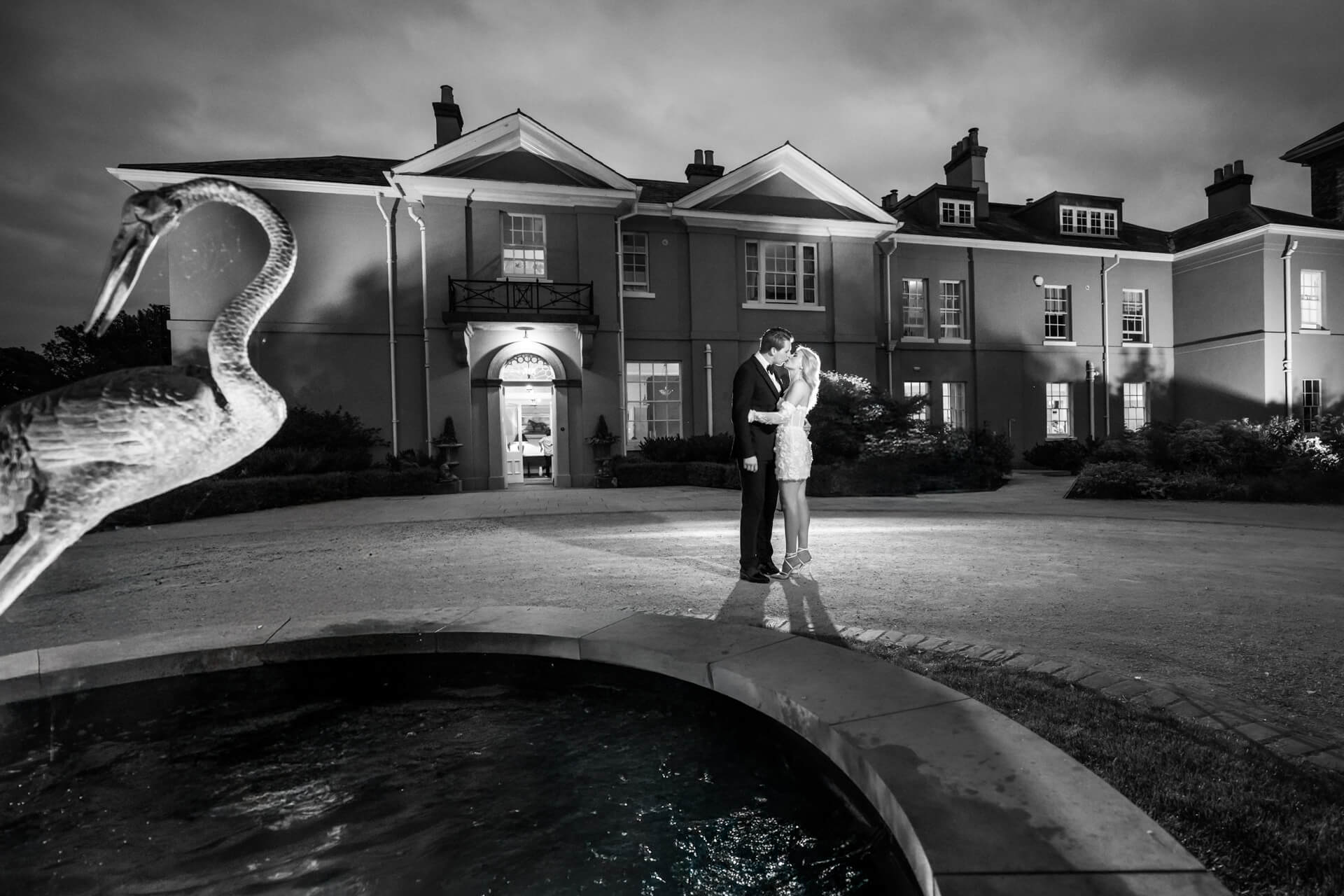 A couple embraces in front of a large house at night, illuminated by a spotlight, with a fountain and bird statue in the foreground. Image is in black and white.