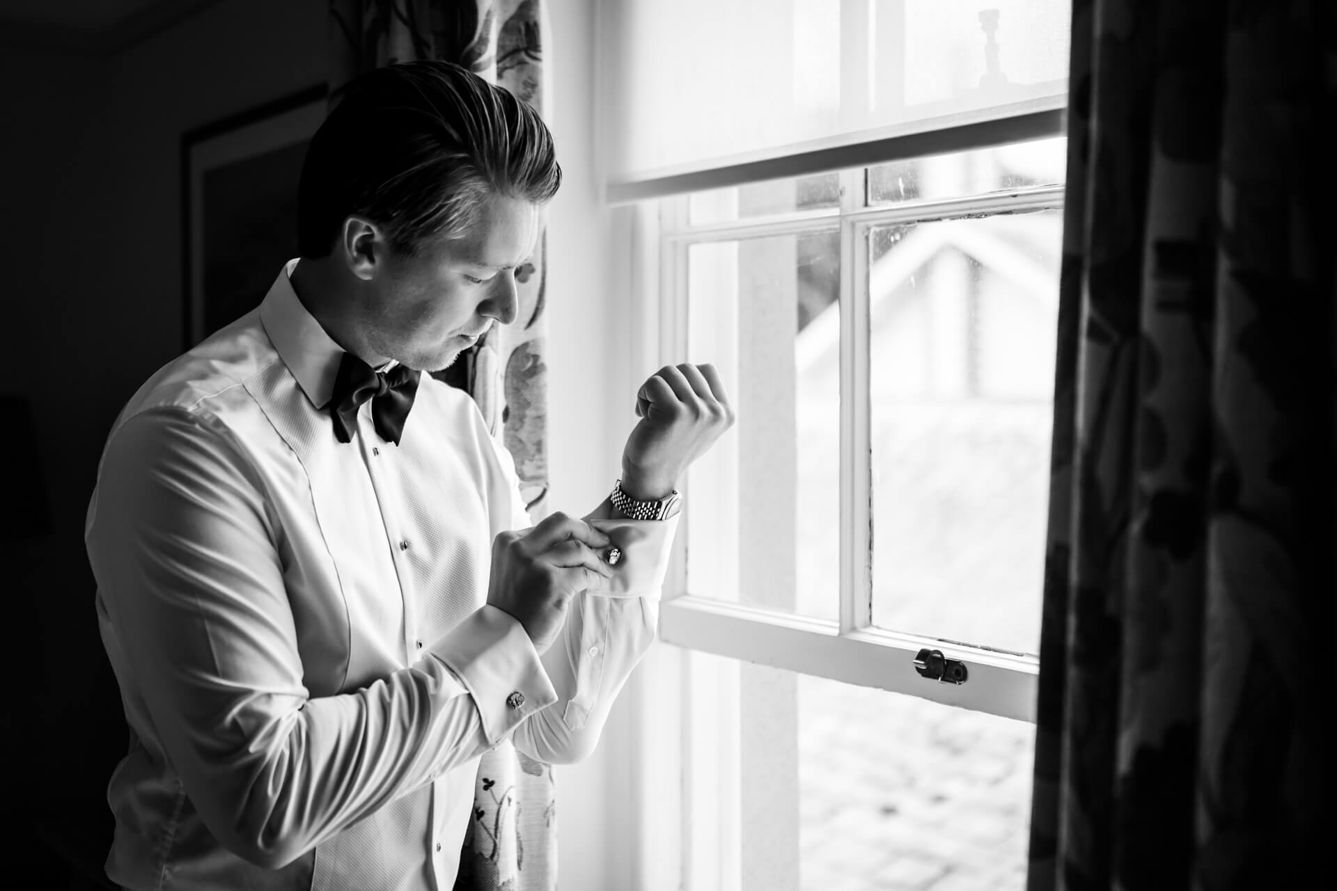 A groom adjusts his watch whilst standing by a window, with natural light illuminating the scene.