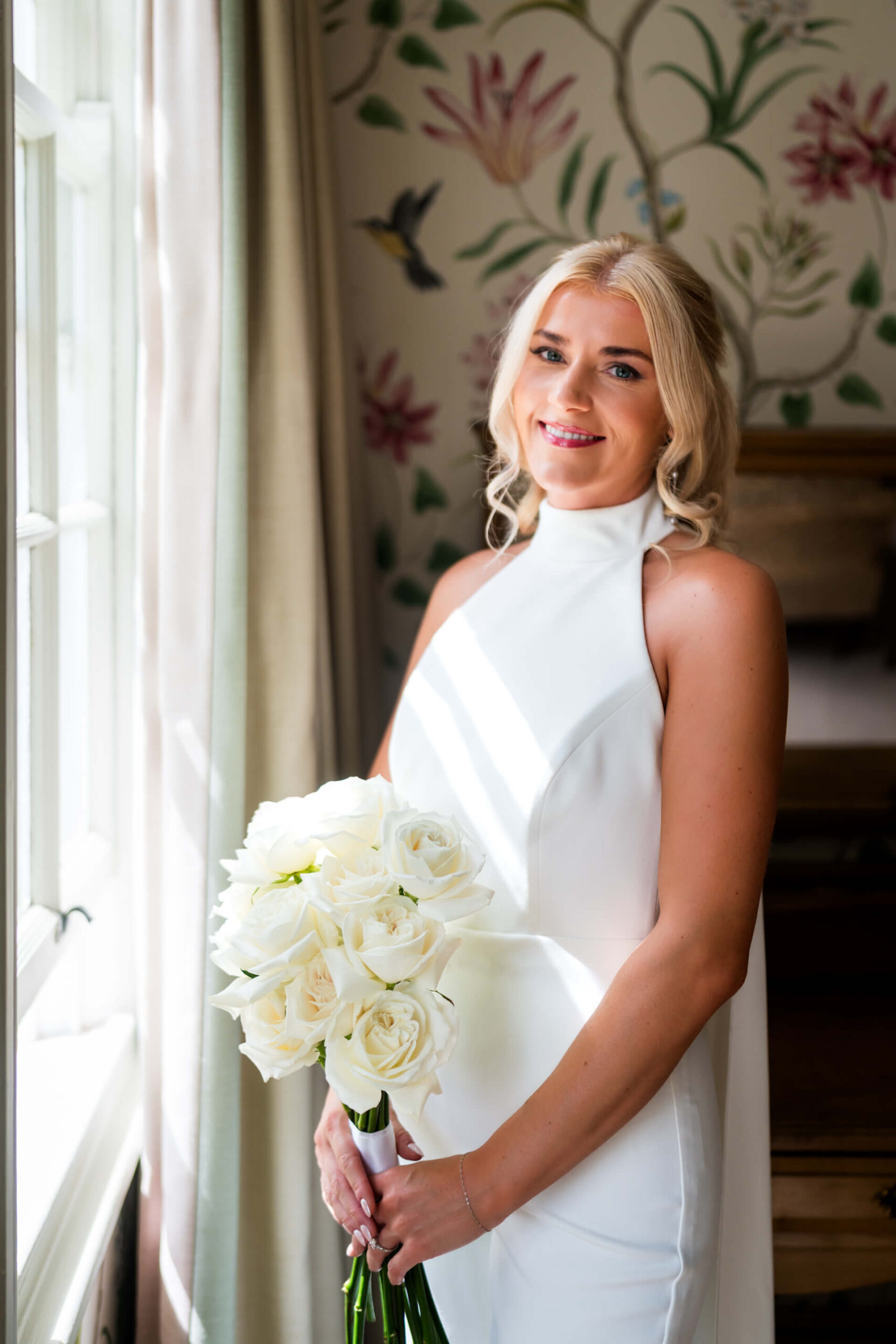 A bride stands by a window holding a bunch of white roses, with light streaming in and floral wallpaper in the background.