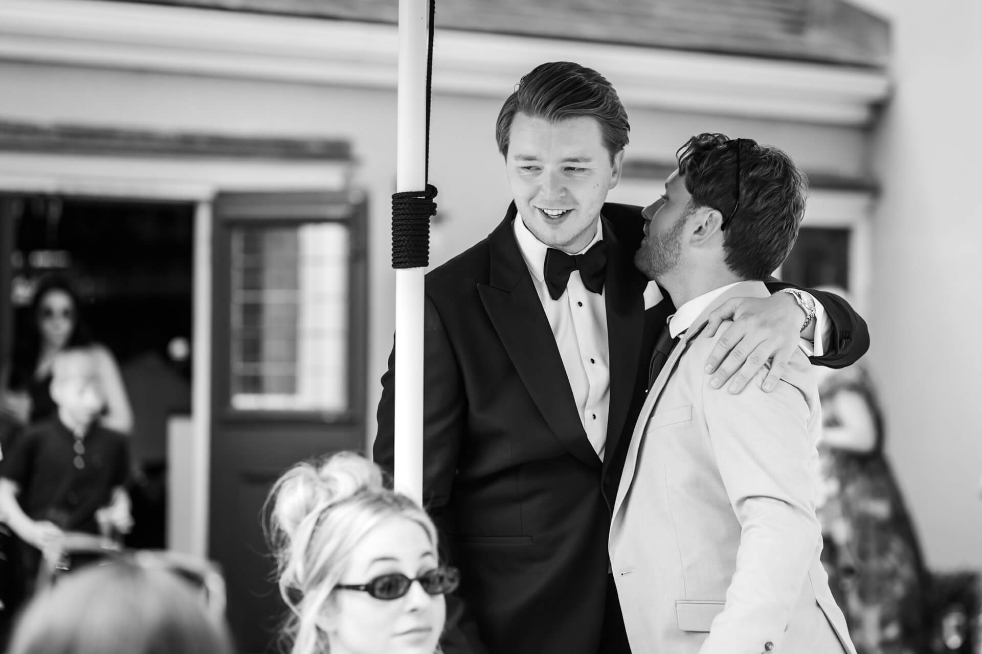 A groom greets his friend at an outdoor wedding gathering